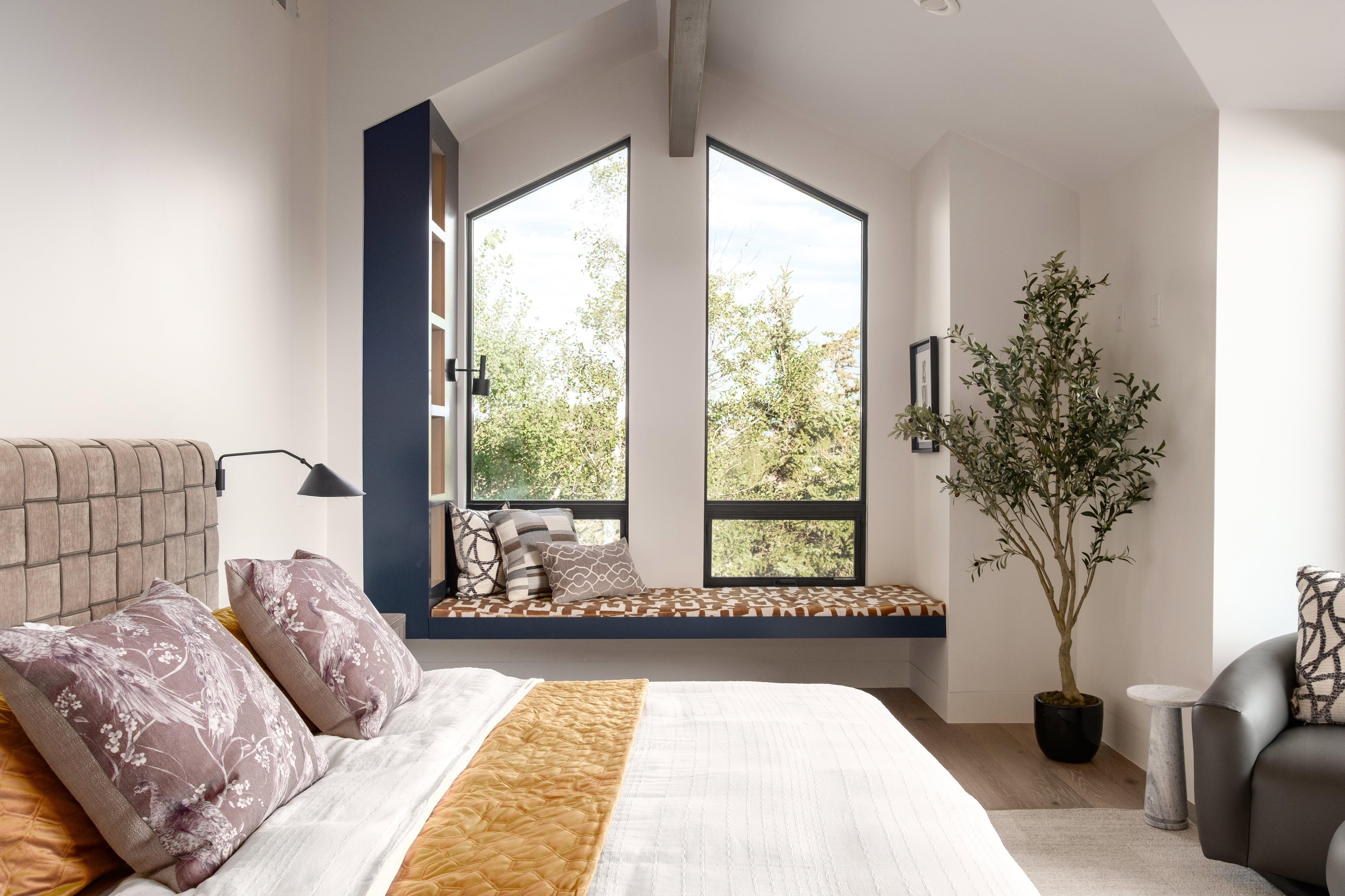 Angled ceiling with exposed beam, two tall triangular windows, built-in window seat, integrated shelving unit, and wood-finish flooring