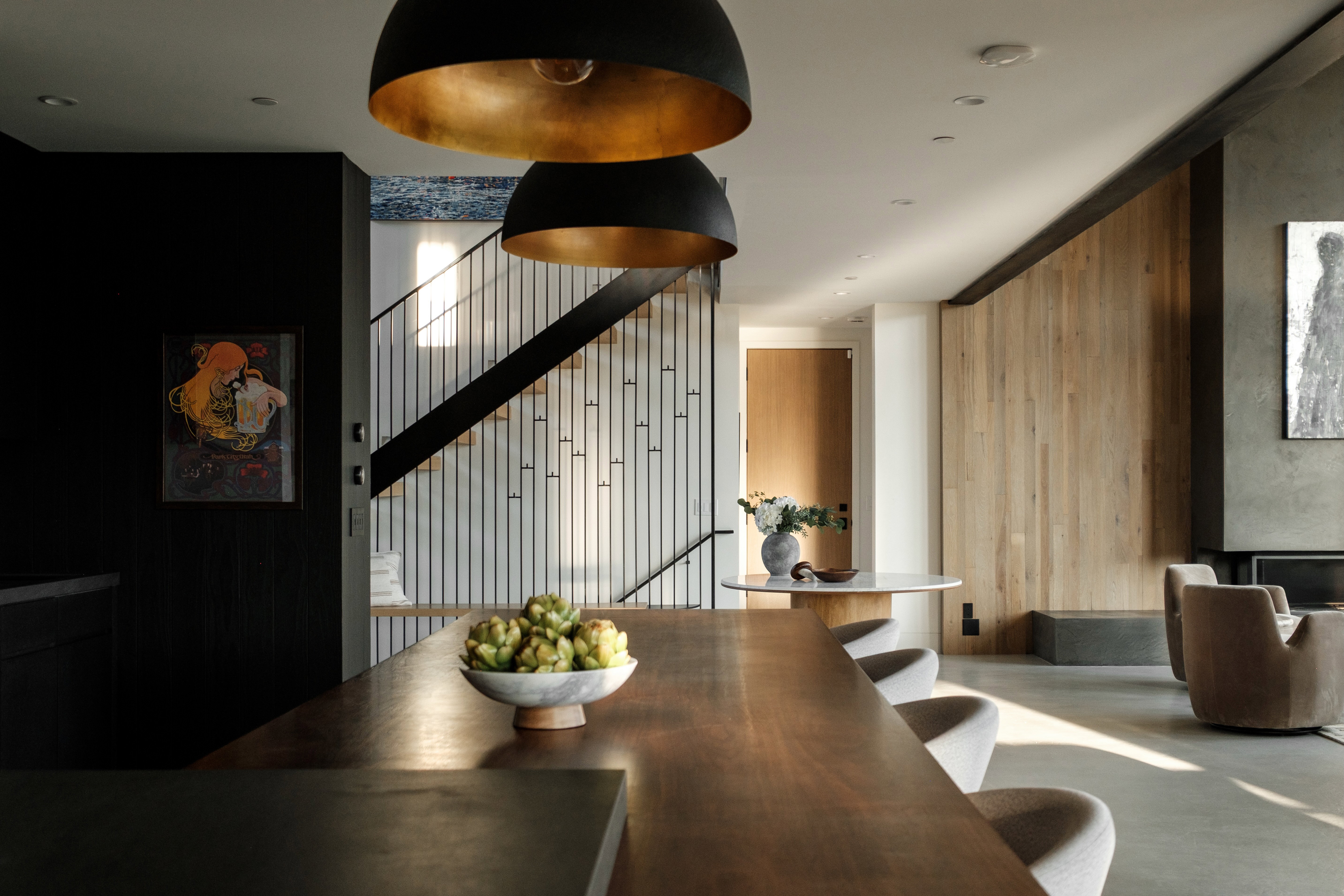 Open-concept space featuring a wood-finish island, two oversized black and gold pendant lights, a staircase with dark metal handrails, a light wood paneled wall, and a concrete-finish fireplace surround