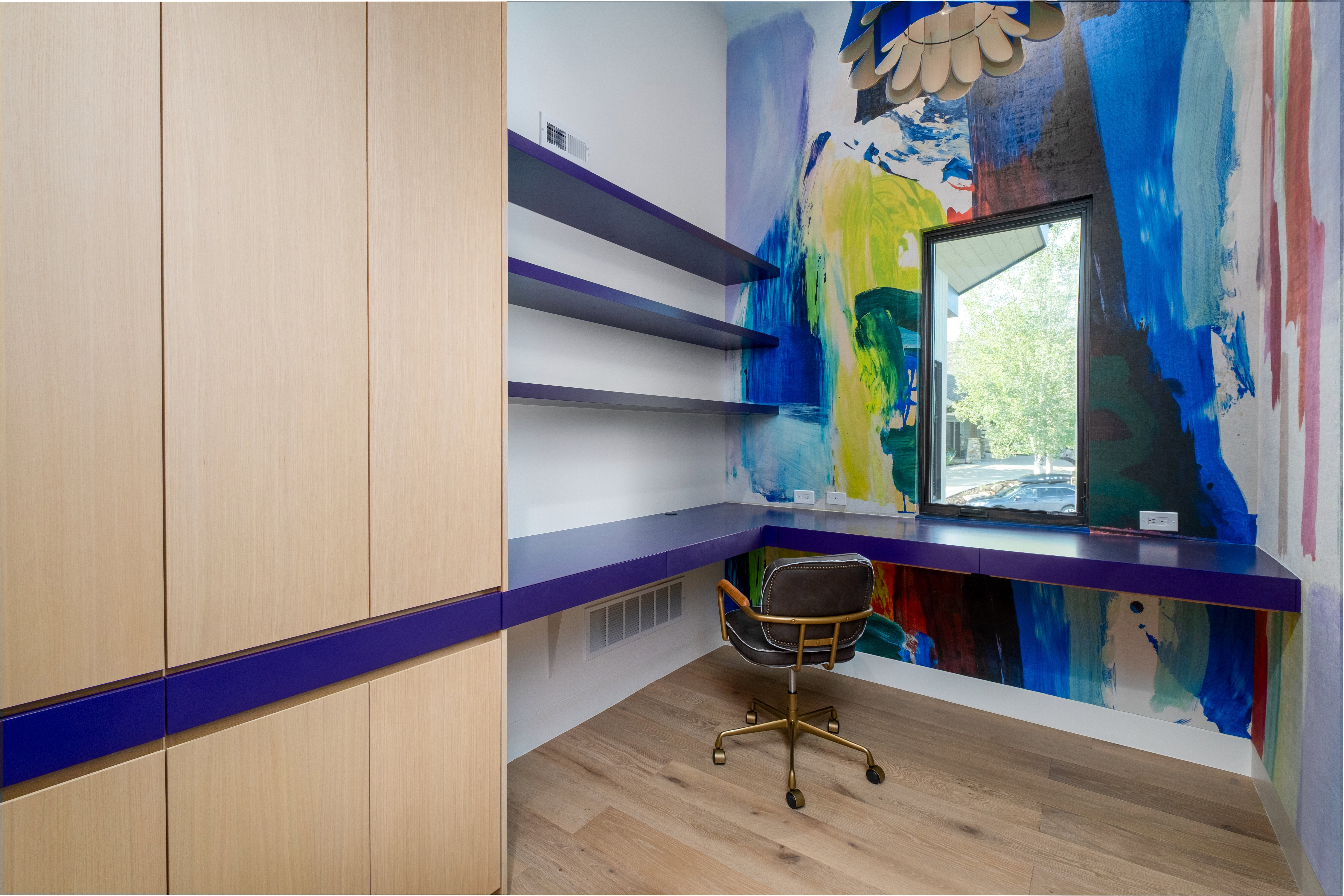 Built-in corner desk with integrated shelving, custom wall covering, large window, wood-finish flooring, and light wood built-in cabinetry