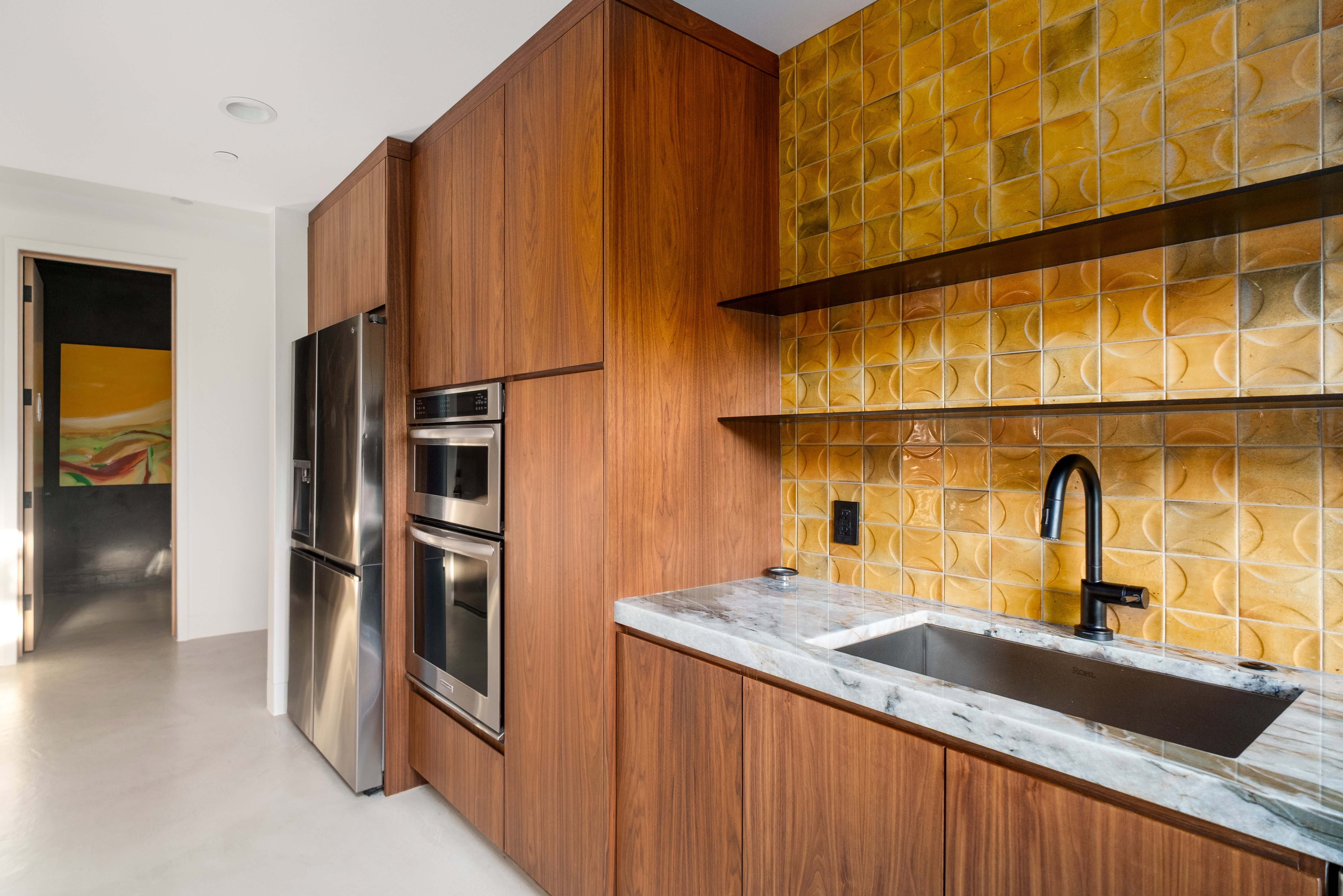 Built-in stainless steel appliances, wood-finish cabinetry, and a textured tile backsplash with open shelving above a sink with a stone countertop