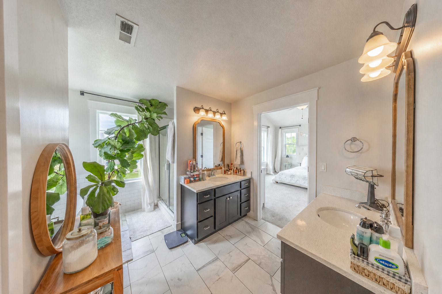 Spacious bathroom featuring dual vanities with undermount sinks and light-toned countertops, a glass-enclosed shower with white subway tile, and a window providing natural light