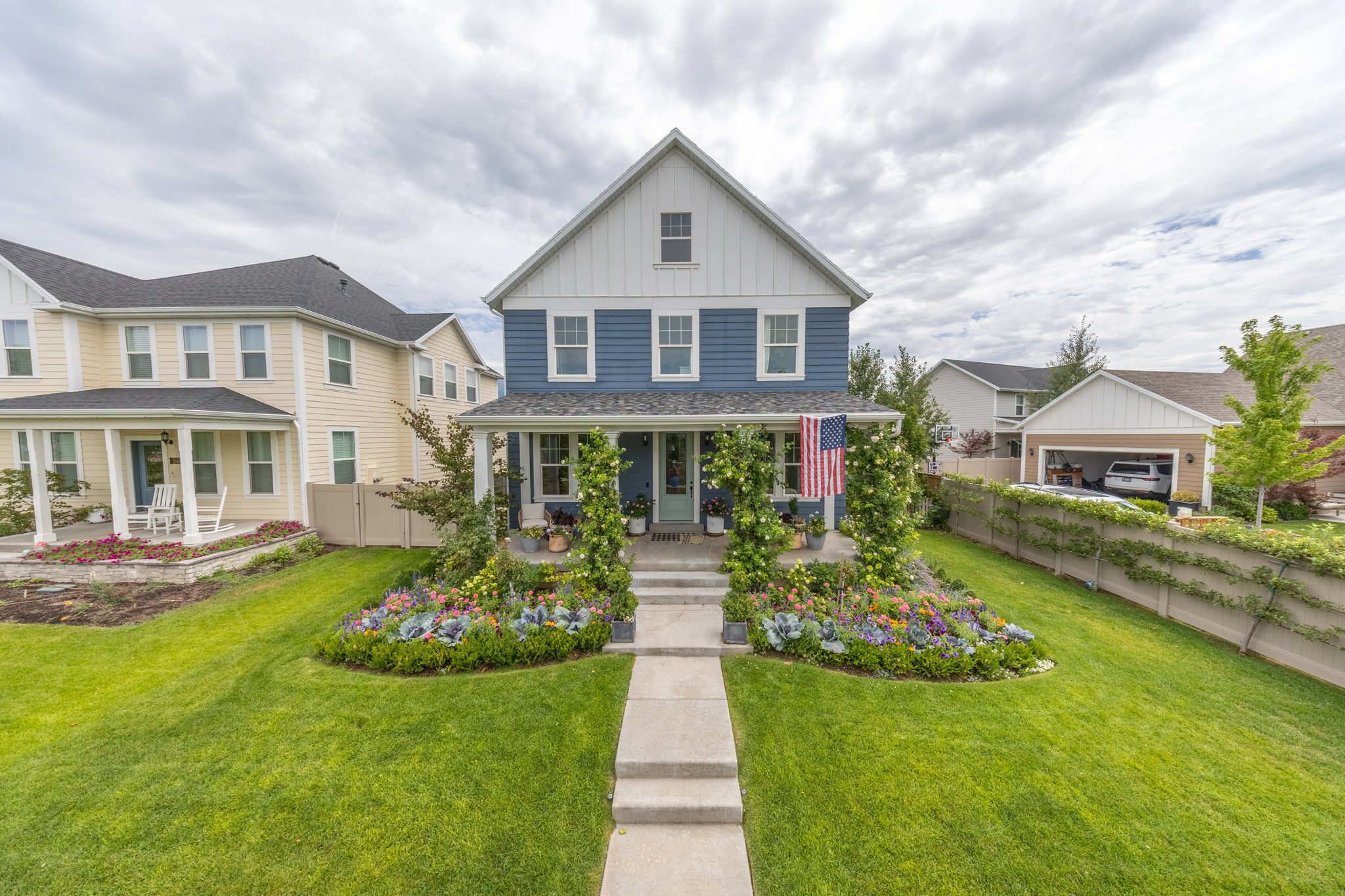 Two-story blue and white siding residence featuring a covered front porch, a well-manicured lawn, a concrete walkway, extensive front garden beds, and a detached garage structure