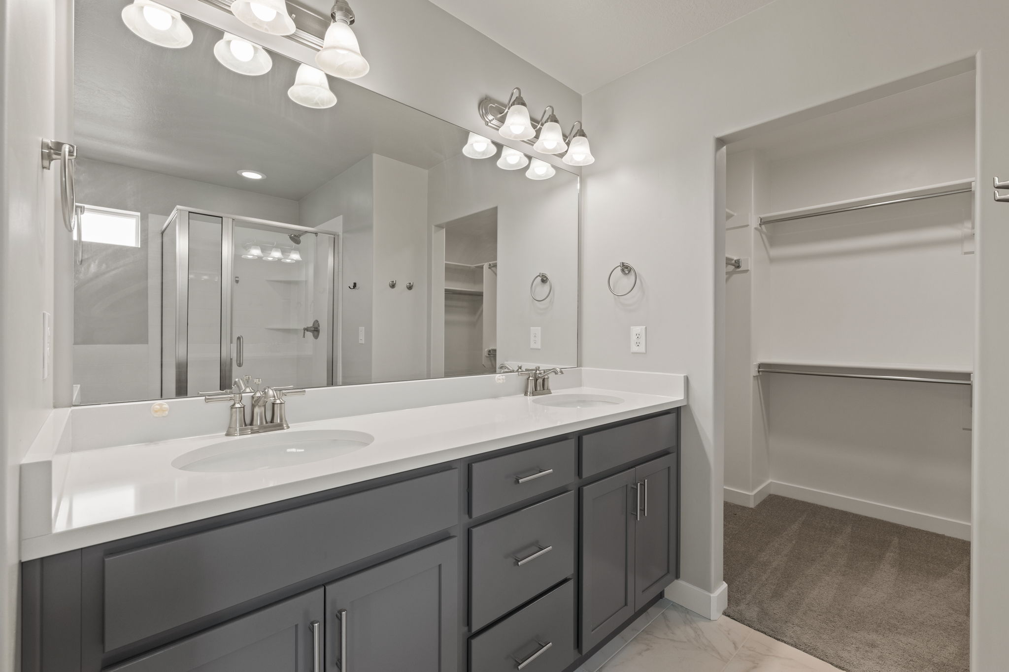 Bathroom featuring a dual vanity with a white countertop and gray cabinetry, a large wall-mounted mirror, a glass-enclosed shower, and a walk-in closet with built-in shelving and hanging rods