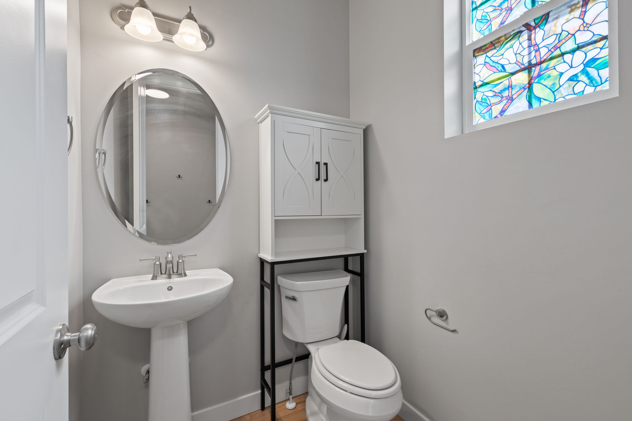 Pedestal sink with a brushed nickel faucet, oval mirror, and a stained-glass window