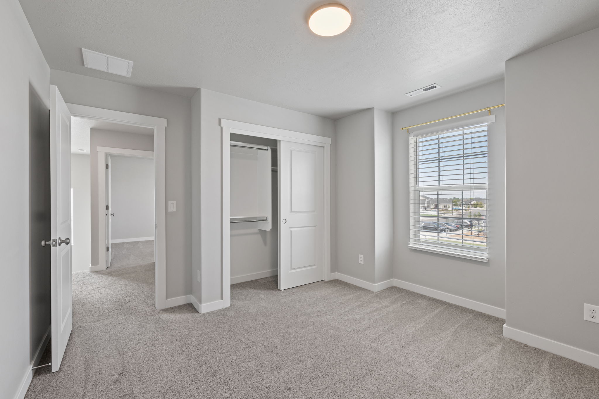 Spacious room featuring light gray carpet, neutral wall paint, a double-door closet with shelving, a window with horizontal blinds, and overhead flush mount lighting