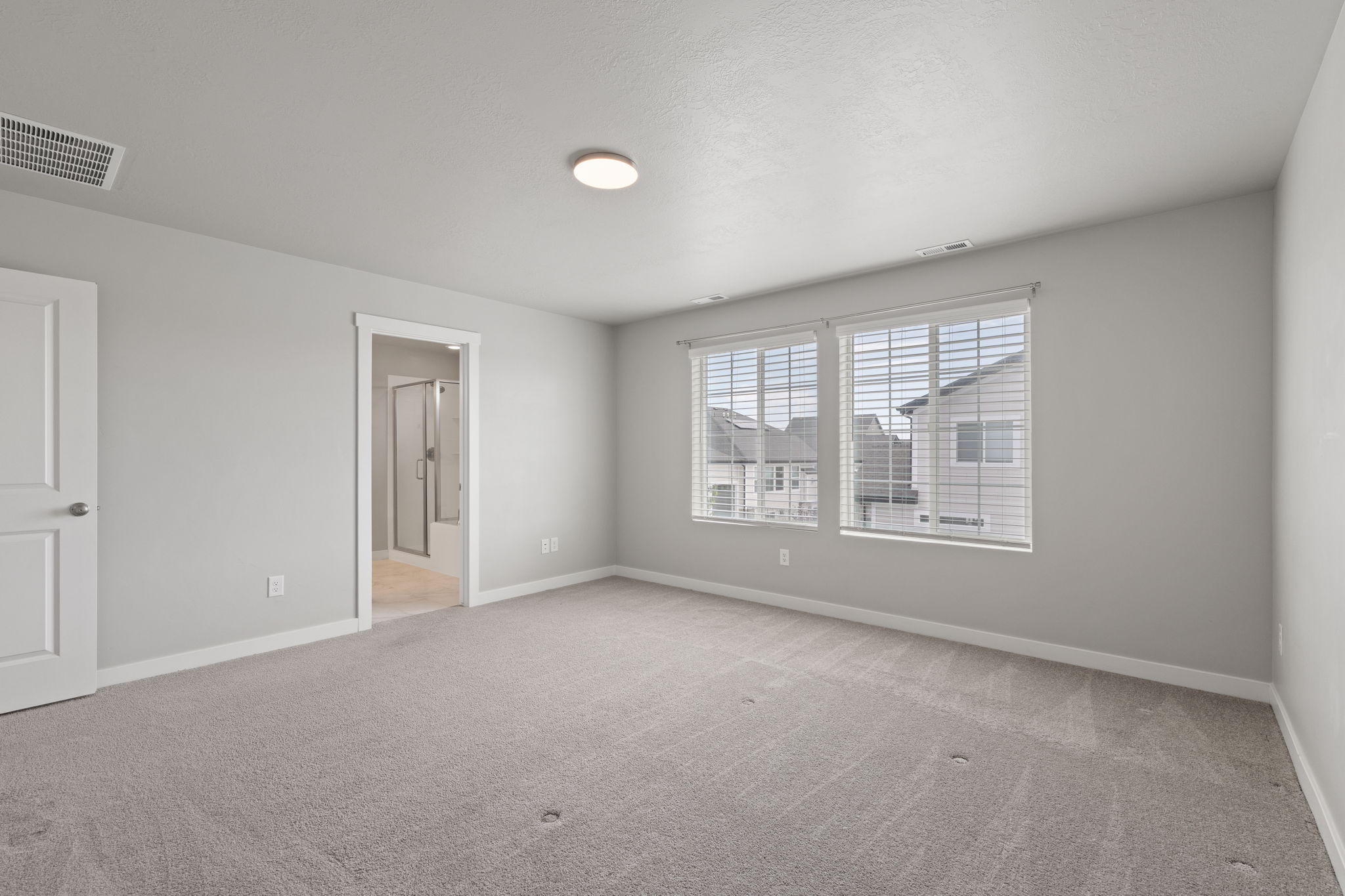 Carpeted room with a ceiling-mounted light fixture, dual windows with blinds, a white paneled door, and an entryway featuring a bathroom with a shower