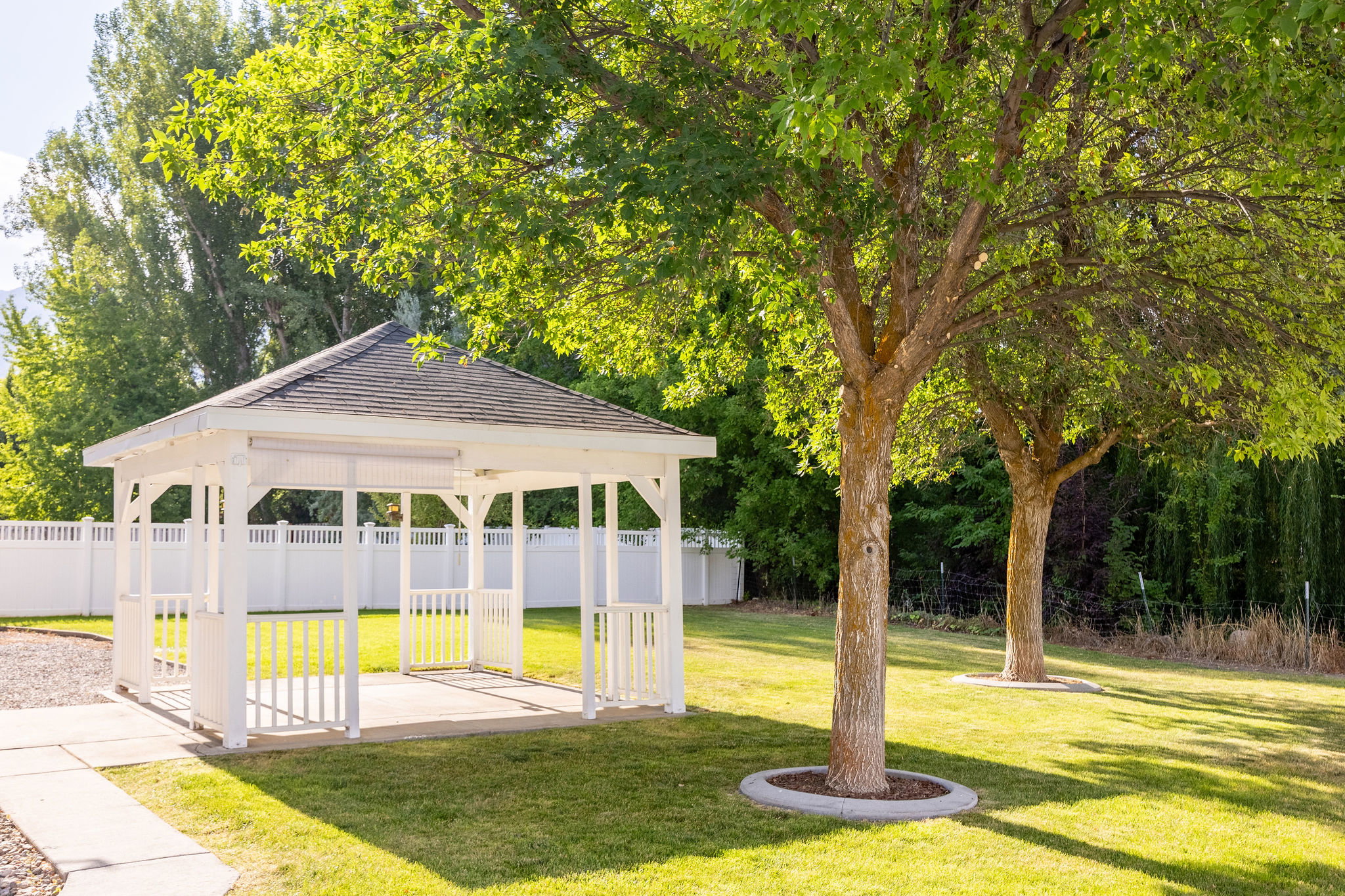 White gazebo with a dark shingled roof, set on a concrete pad with an adjacent paved pathway