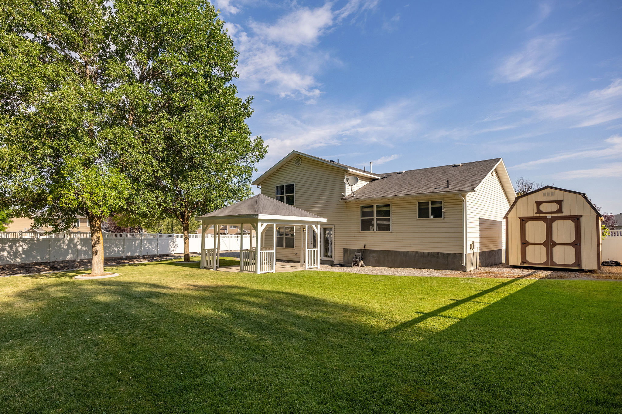 Spacious rear yard featuring an expansive lawn, mature shade trees, a covered patio with a white gazebo, a storage shed, and a privacy fence