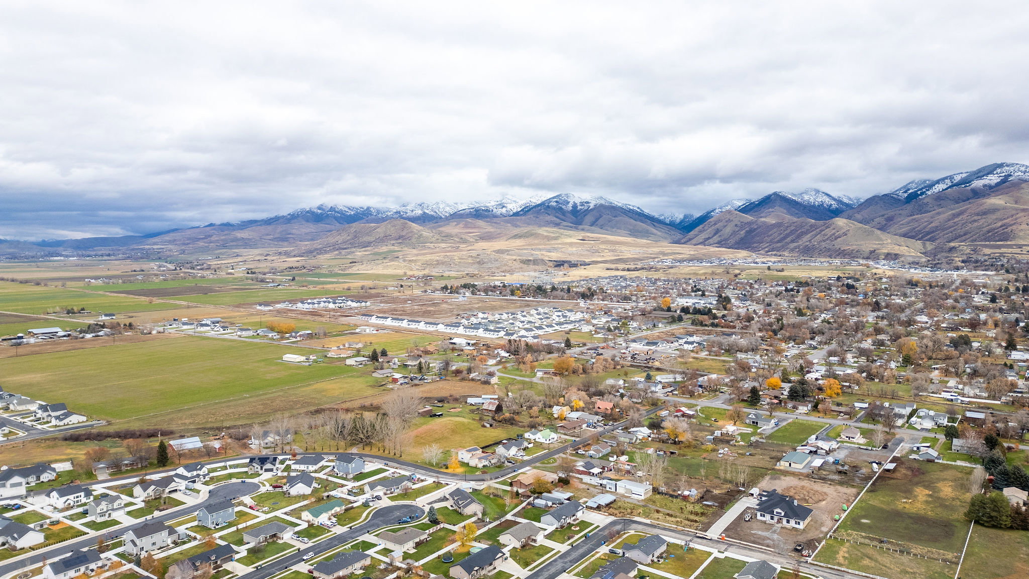 Aerial perspective showcasing an expansive residential area with varied housing styles, surrounded by open fields and distant snow-capped mountains