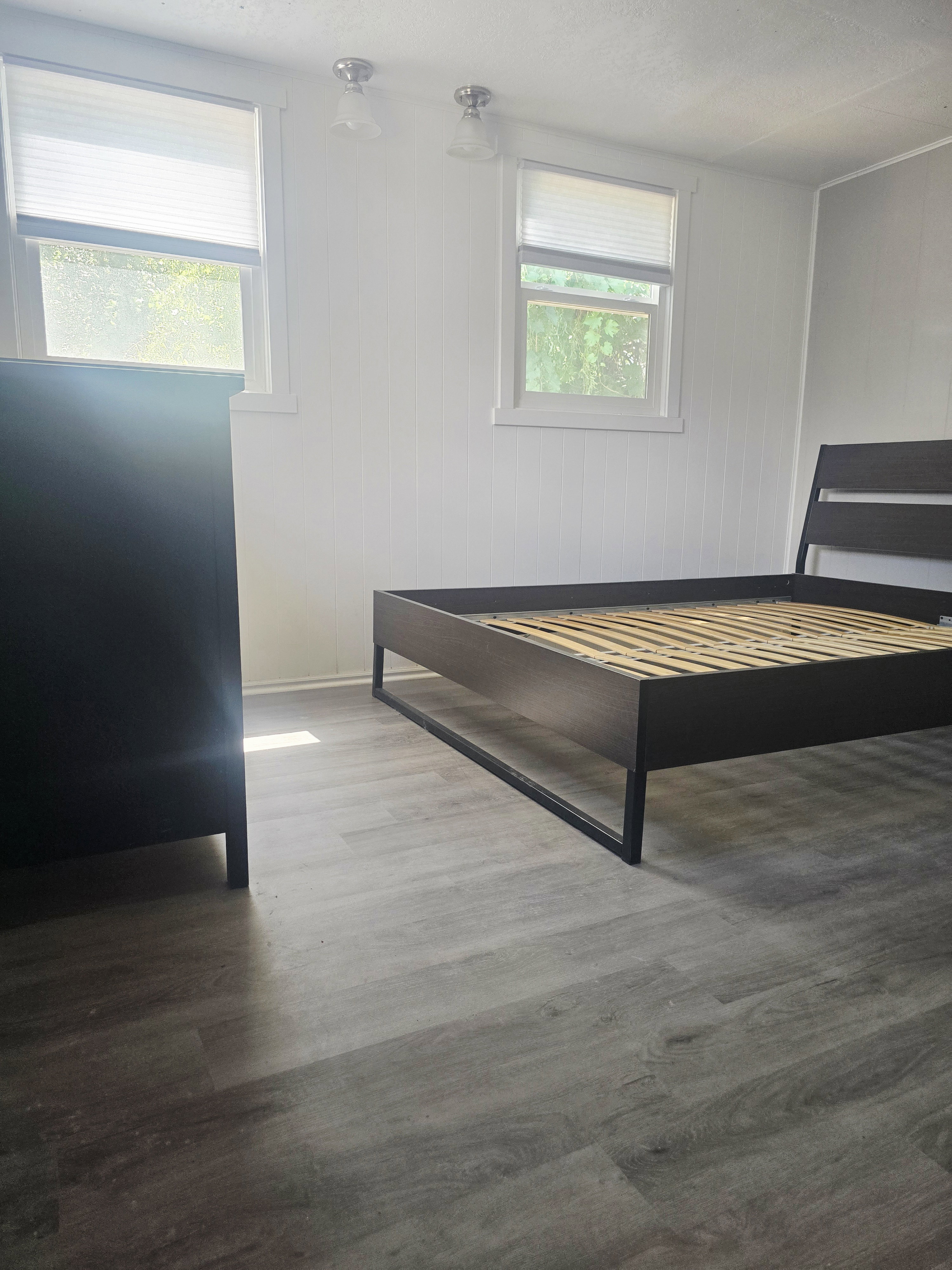 Room featuring gray wood-finish flooring, two windows with white blinds, white vertical paneling on walls, and two ceiling-mounted light fixtures