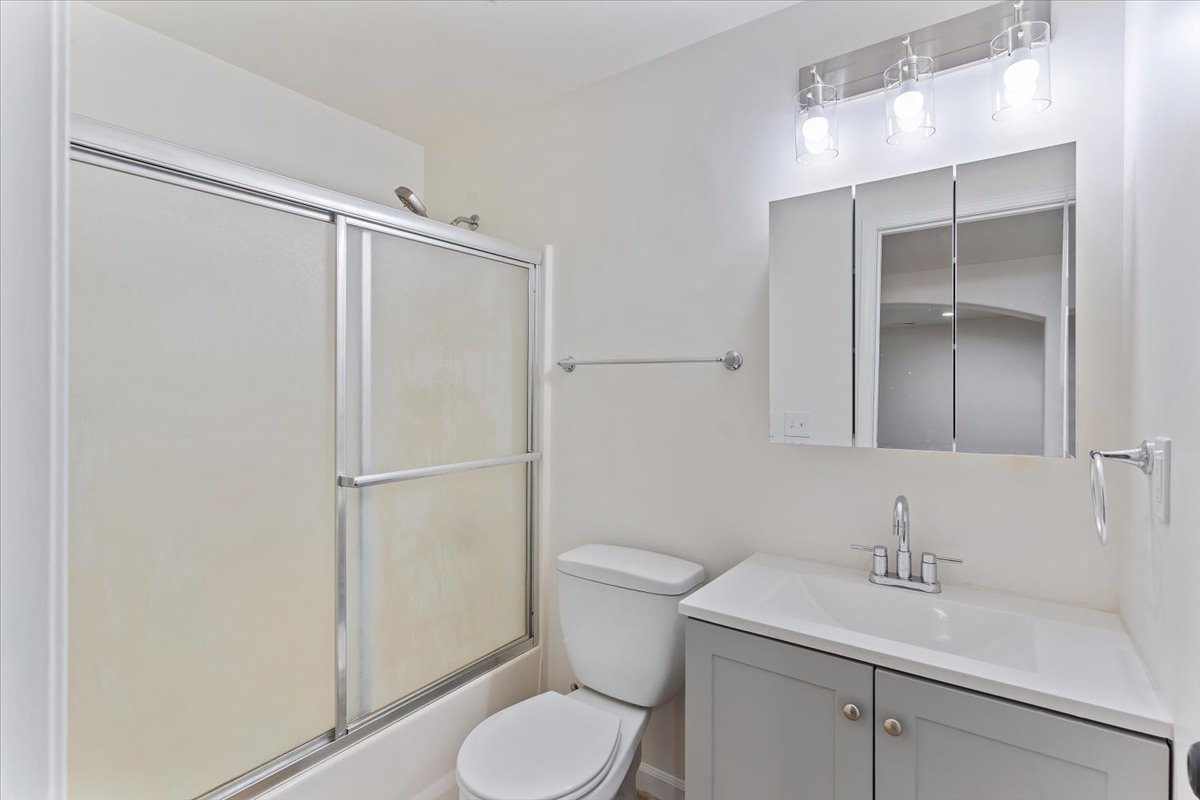 Bathroom featuring a white bathtub with frosted sliding shower doors, a commode, and a vanity with a white countertop and gray cabinetry