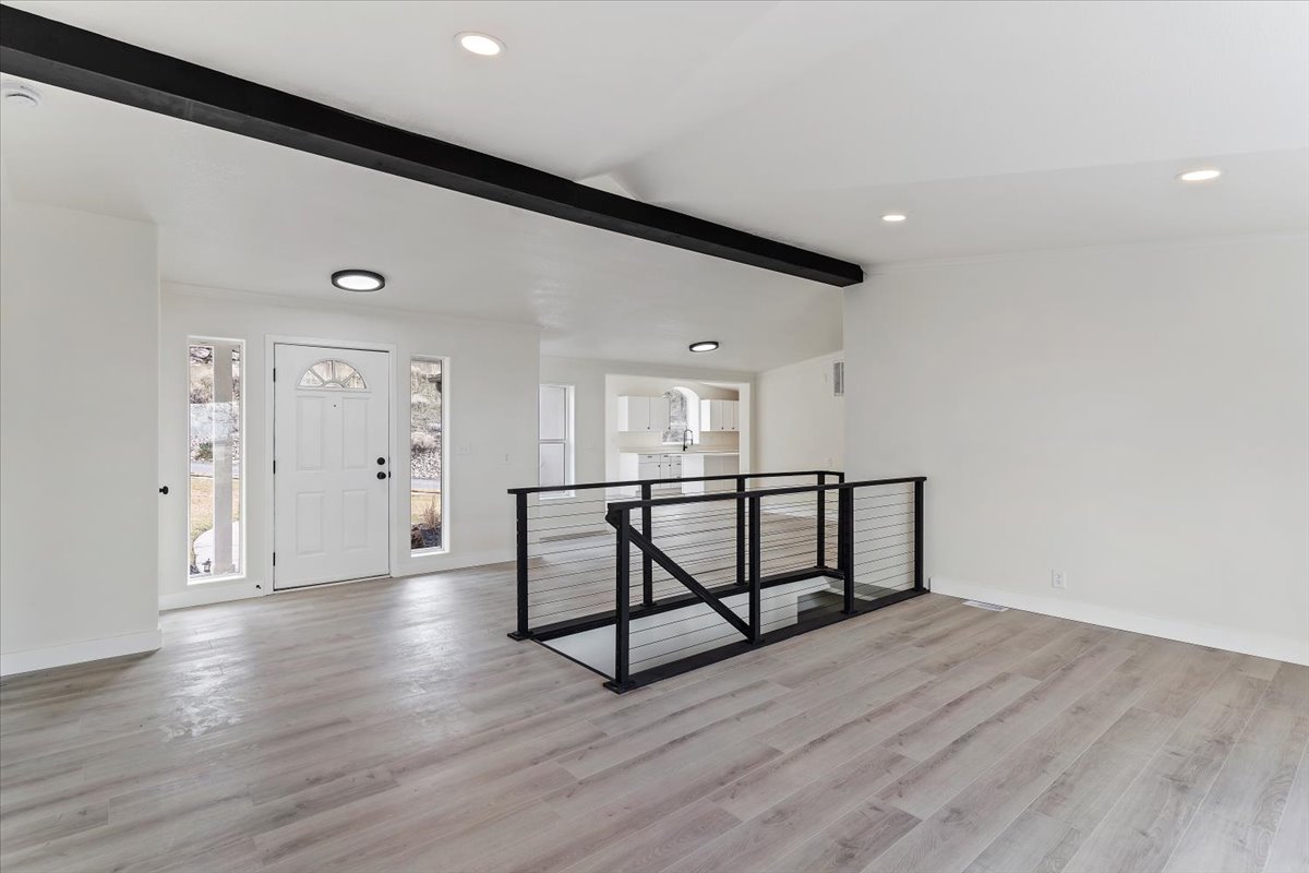 Spacious open-concept living area featuring light wood-finish flooring, a modern cable railing system, and a prominent black structural beam