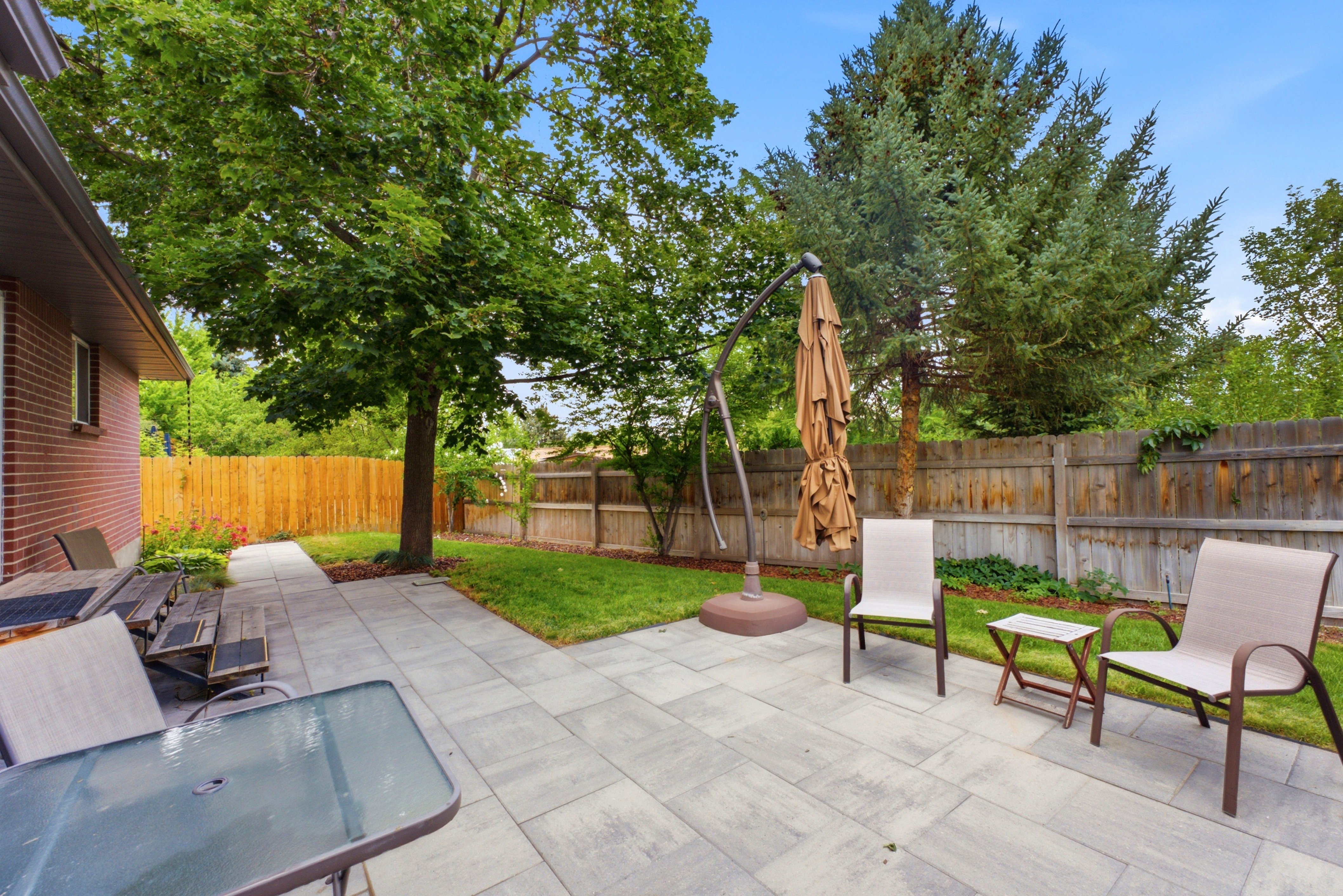 Expansive backyard featuring a large paver patio, brick exterior wall, mature trees, manicured lawn, and fully fenced yard. Perfect for entertaining!