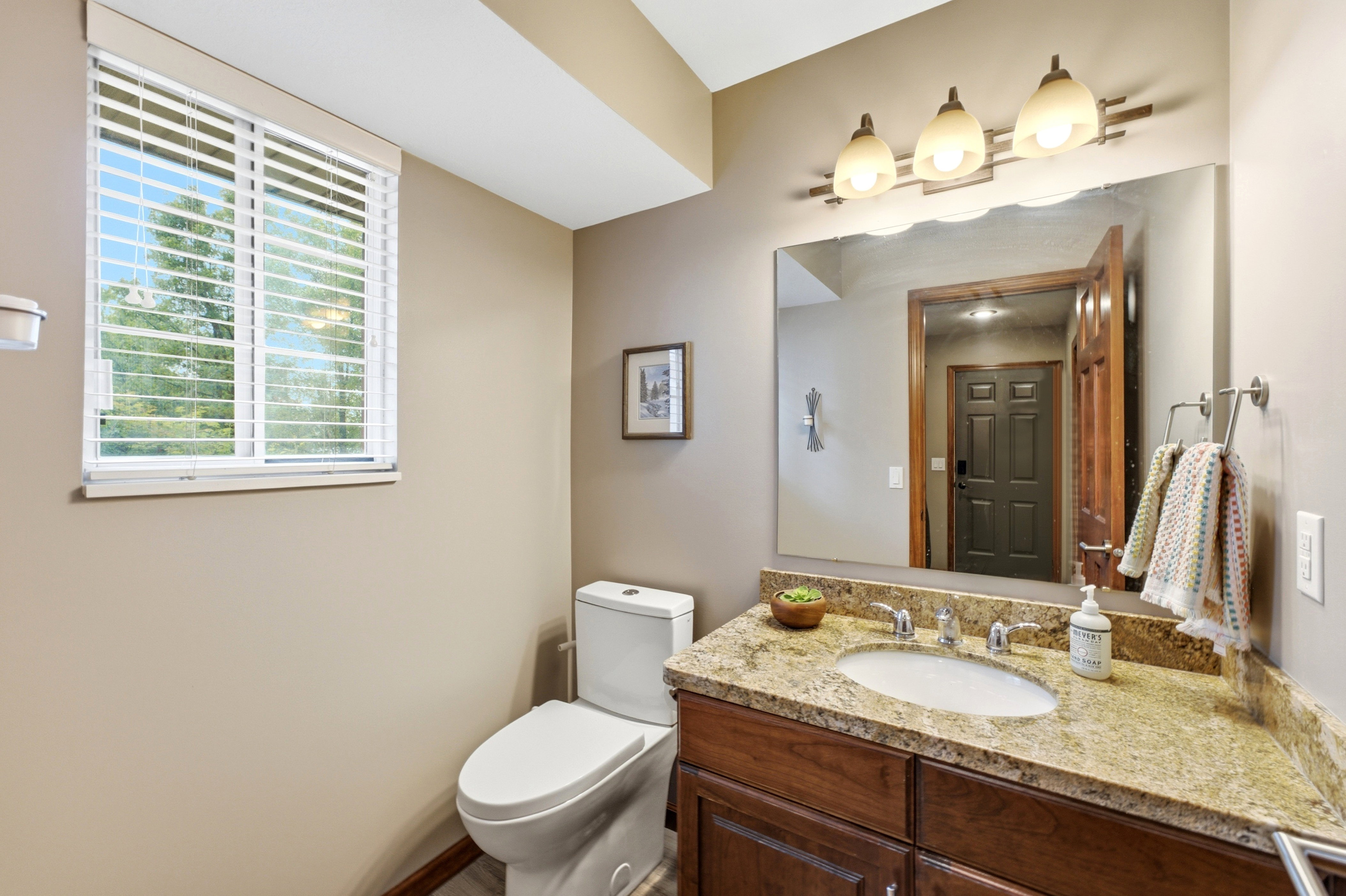 Powder bathroom on the main level featuring a window with white blinds, a toilet, a vanity with a granite-style countertop, an undermount sink, and a framed mirror with a three-light fixture.
