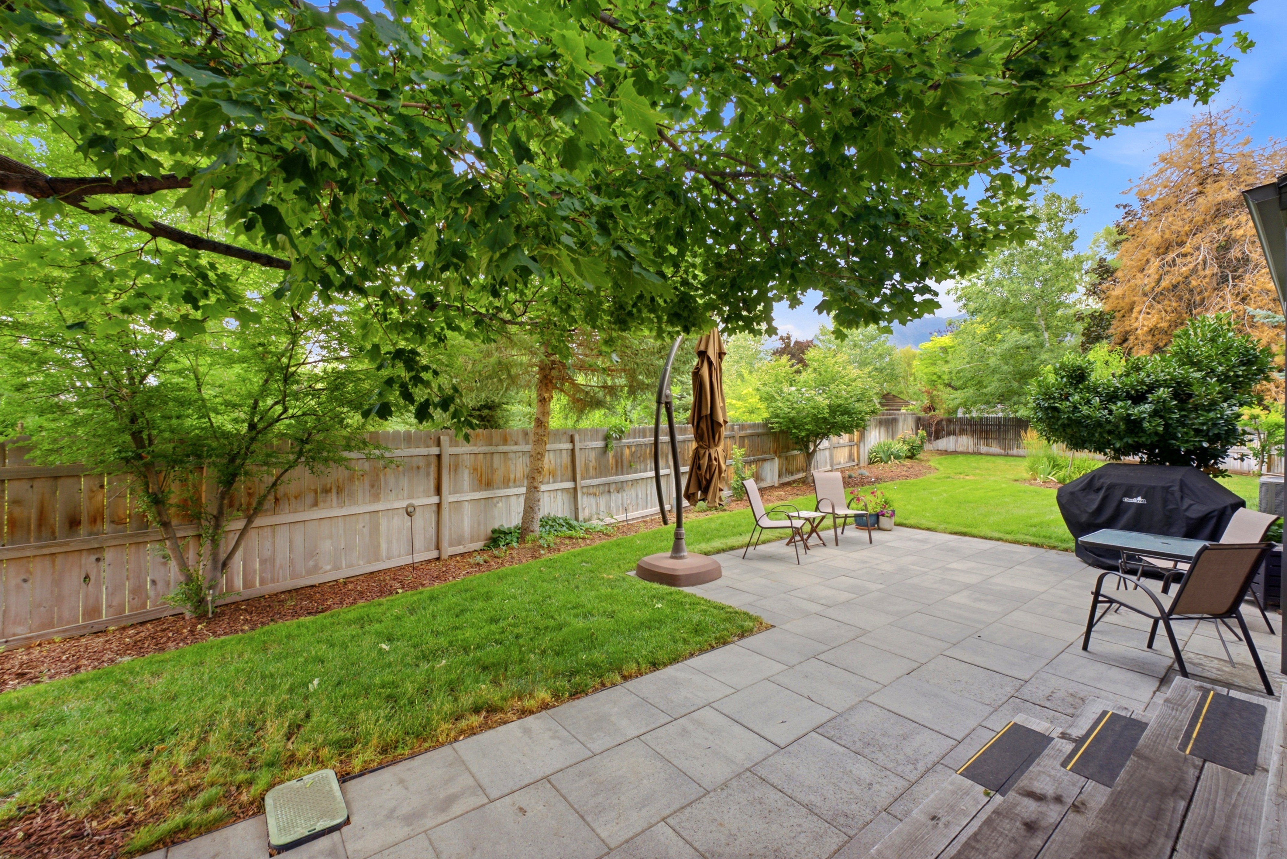 Expansive backyard featuring a large paver patio, brick exterior wall, mature trees, manicured lawn, and fully fenced yard. Perfect for entertaining!