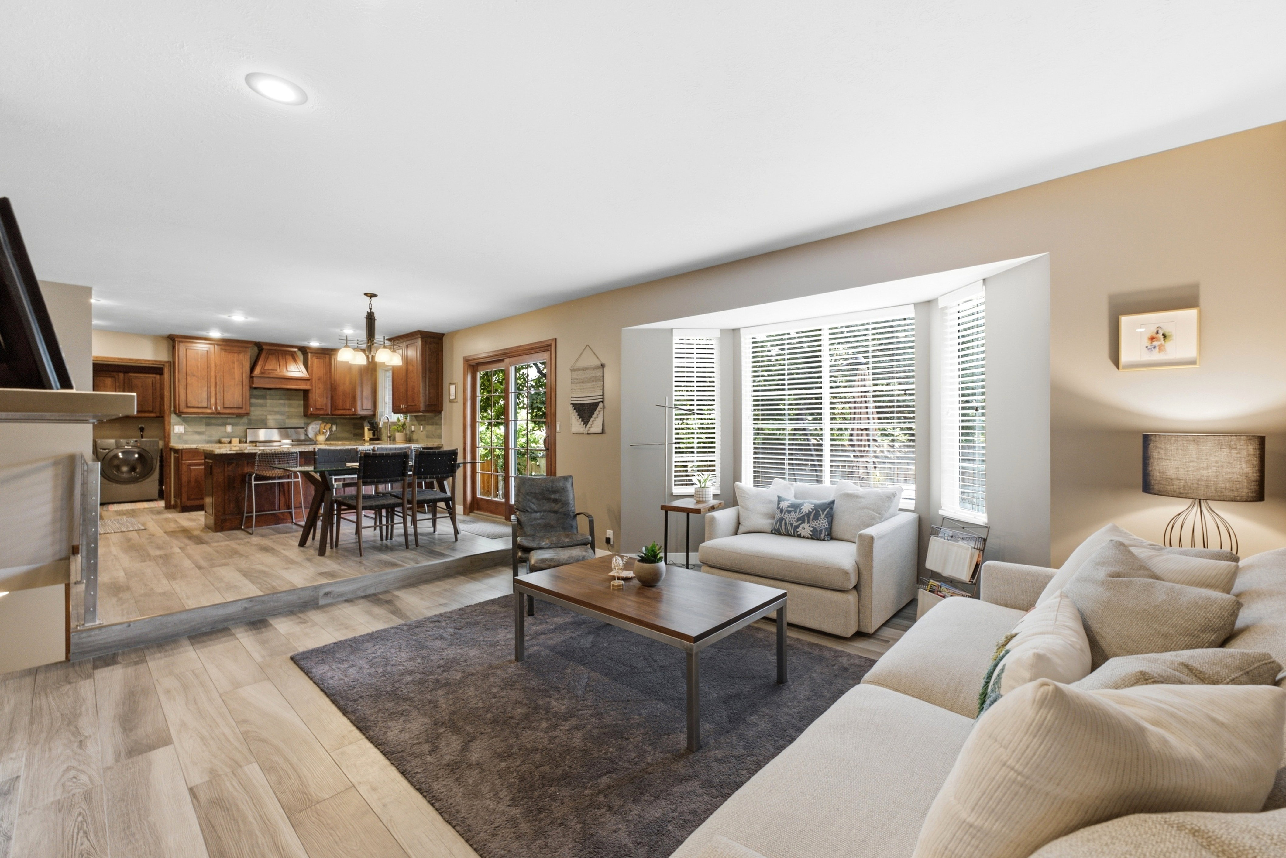 Open concept living space featuring light wood-finish flooring, a bay window with blinds, recessed lighting, and a defined kitchen area with dark wood cabinetry and a sliding glass door.