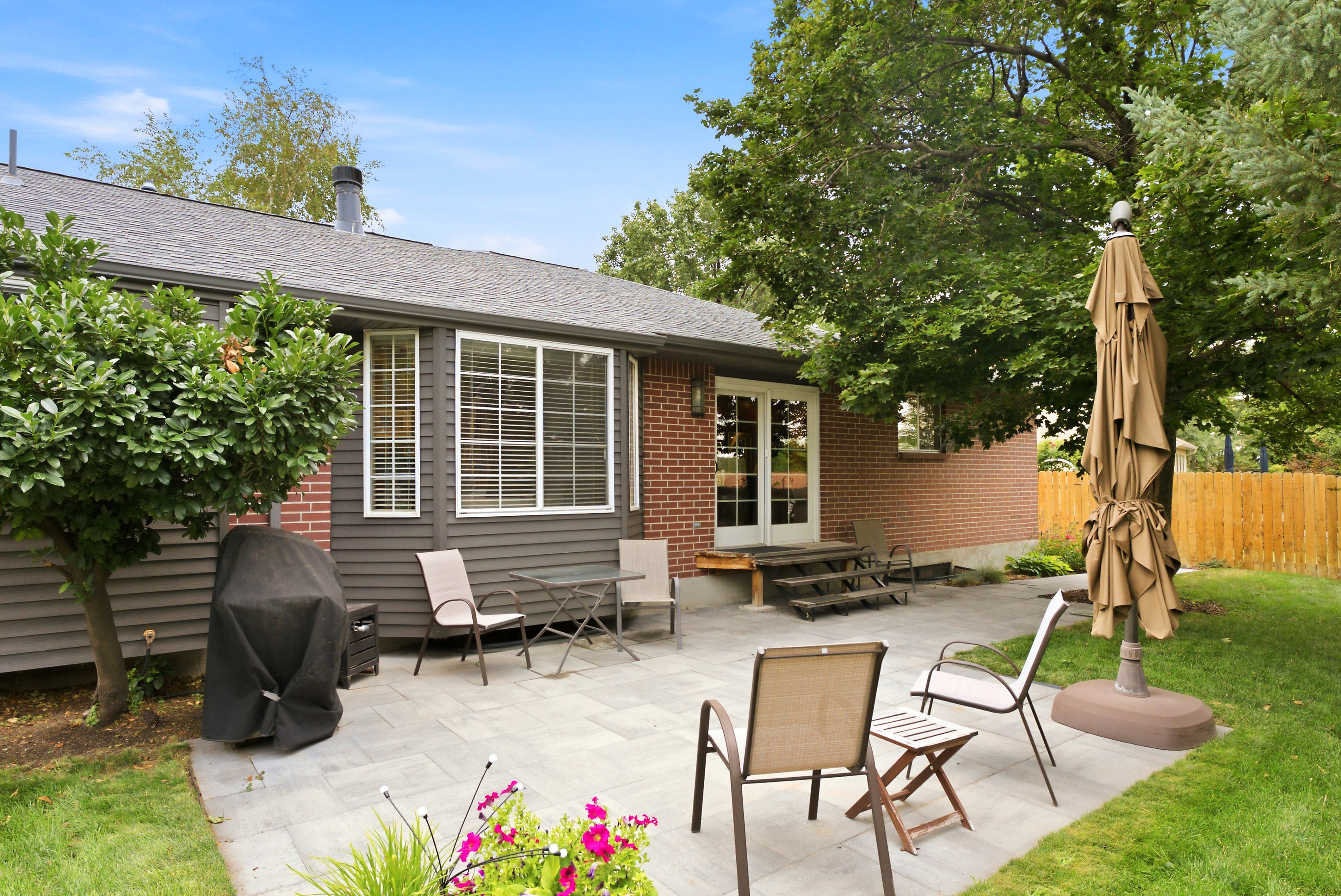 Expansive backyard featuring a large paver patio, brick exterior wall, mature trees, manicured lawn, and fully fenced yard. Perfect for entertaining!