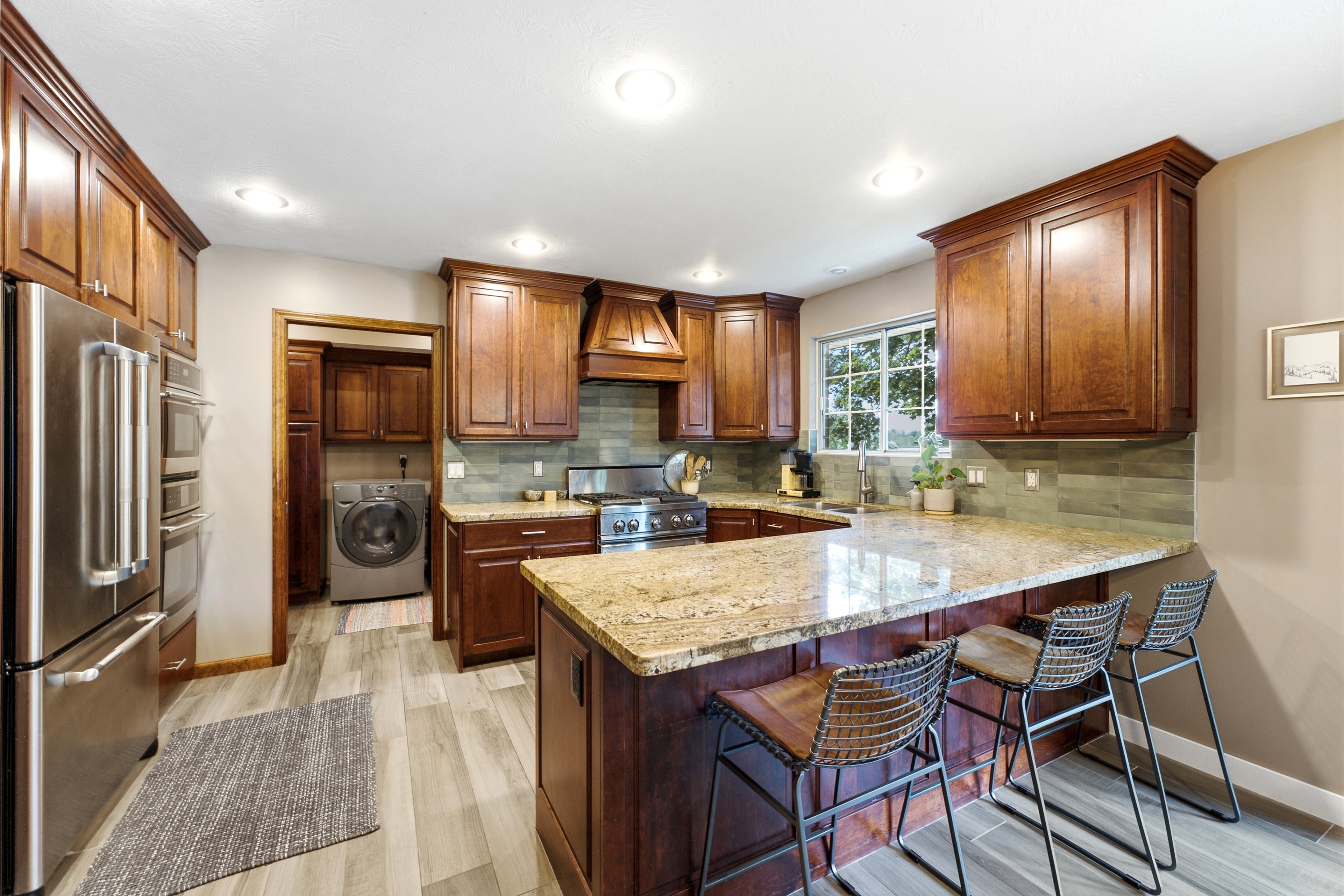 Well designed kitchen featuring rich wood cabinetry, stainless steel appliances, a large island with a stone countertop, a tile backsplash, and light wood-finish flooring.
