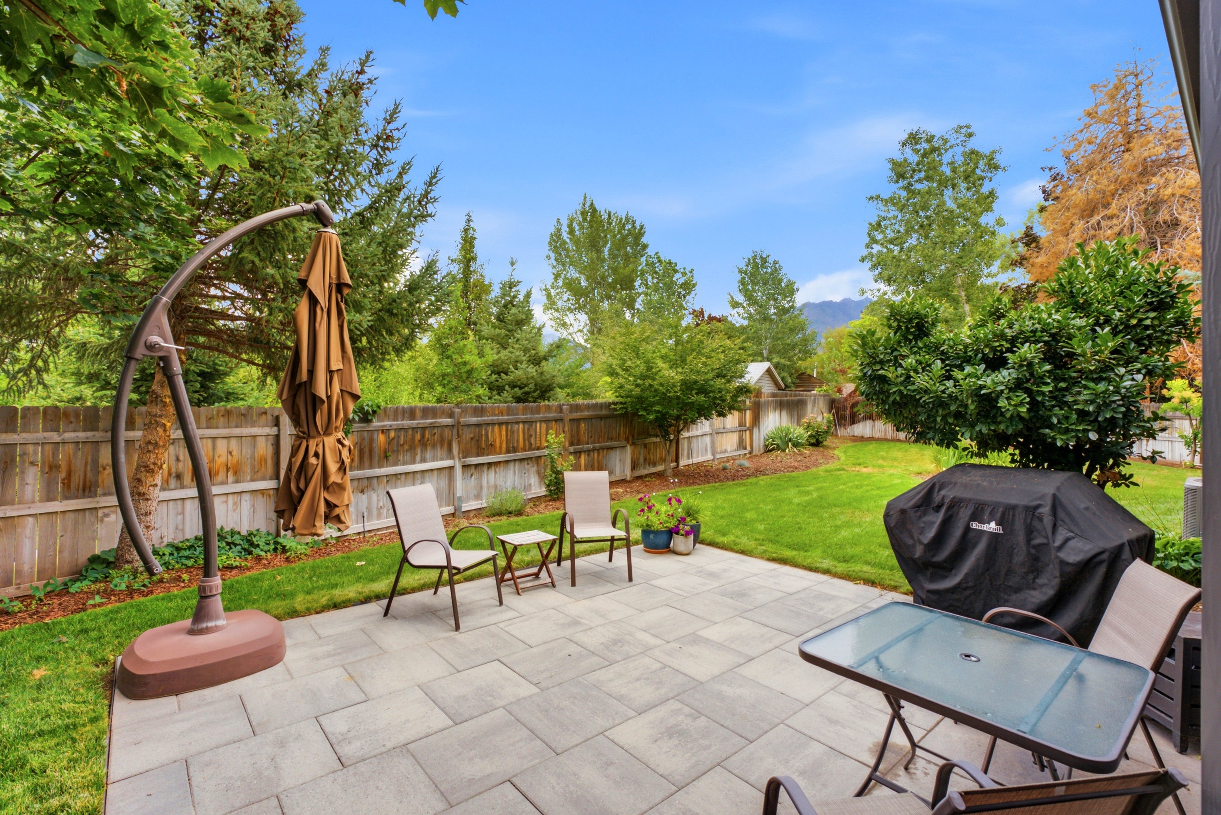Expansive backyard featuring a large paver patio, brick exterior wall, mature trees, manicured lawn, and fully fenced yard. Perfect for entertaining!
