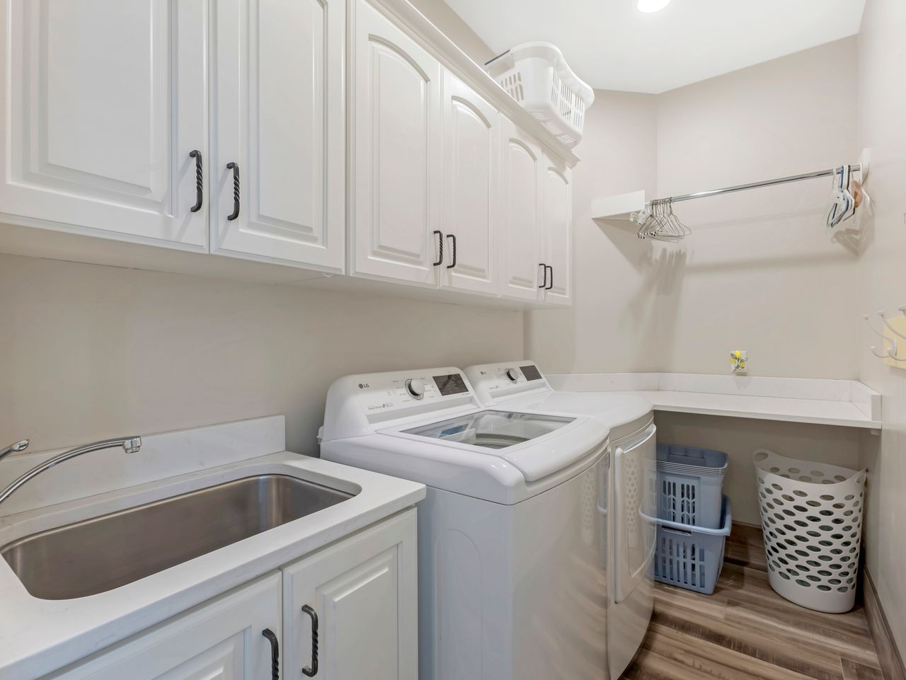 Laundry room featuring white shaker-style cabinetry, a stainless steel utility sink, light-toned countertops, wood-finish flooring, and a chrome hanging rod
