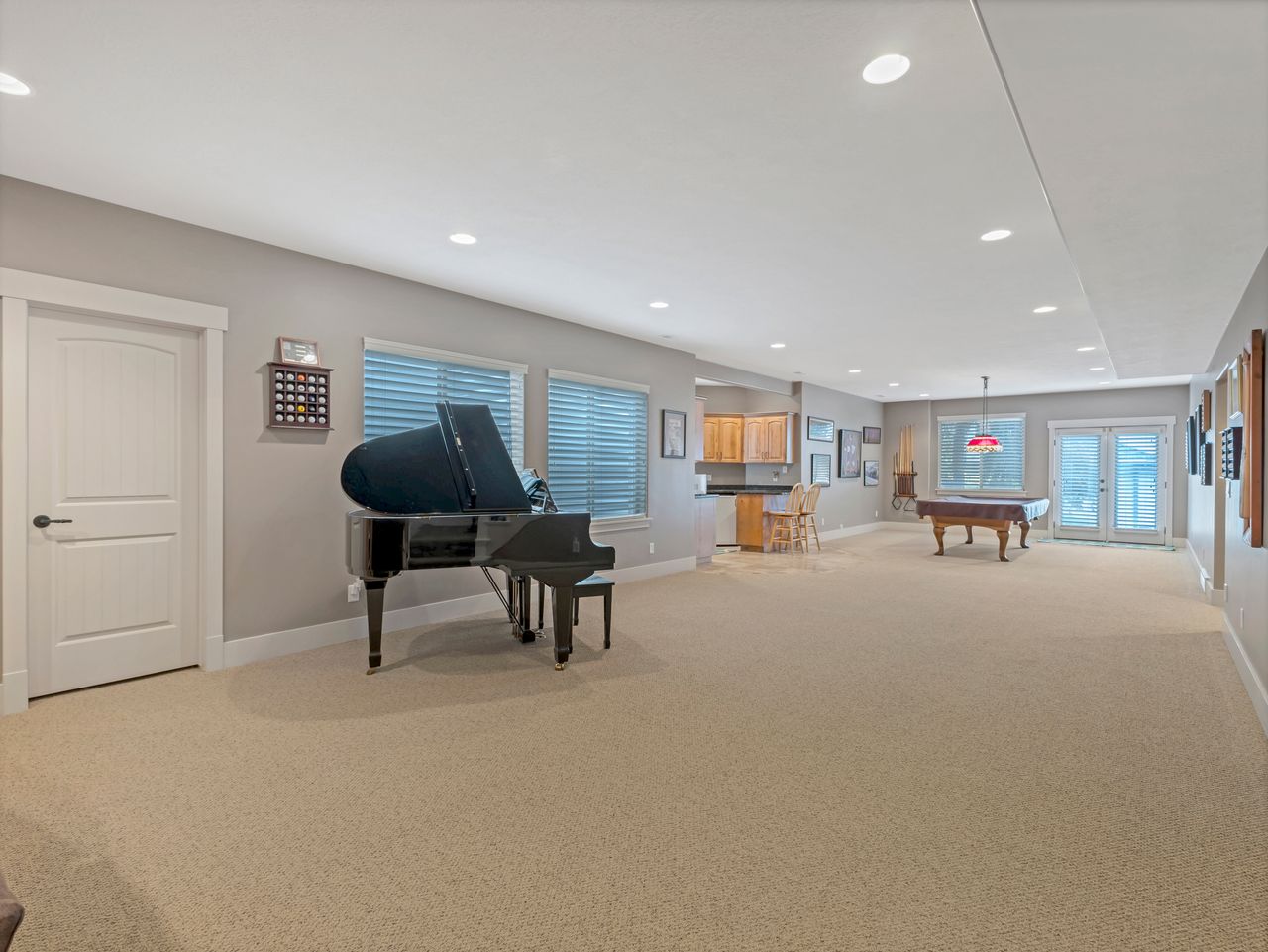 Spacious carpeted room featuring recessed lighting, multiple windows with blinds, a built-in bar with wood cabinetry, and double glass-paneled doors
