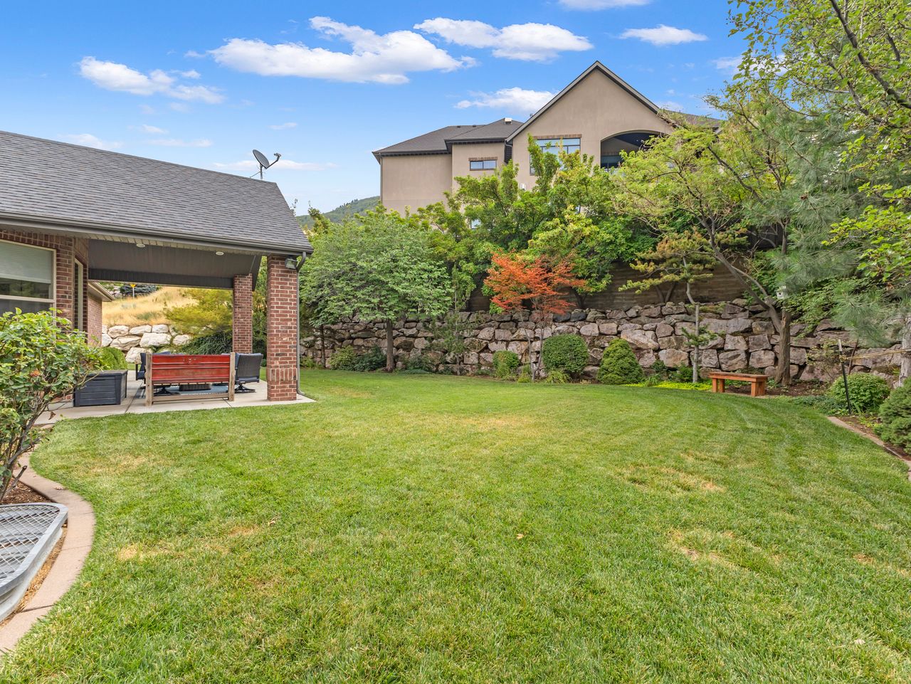 Expansive backyard featuring a manicured lawn, a covered patio with brick columns, and a robust stone retaining wall