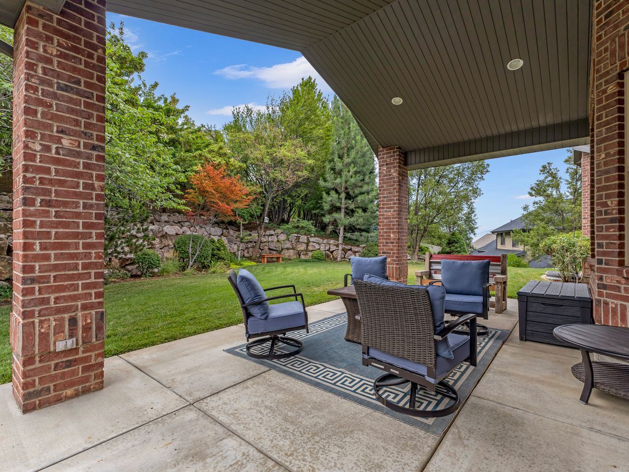 Covered concrete patio featuring brick pillars, an overhead ceiling with recessed lighting, a stone retaining wall, and mature landscaping