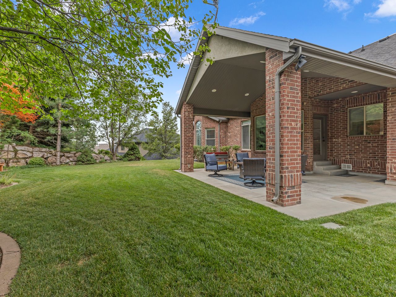 Expansive backyard featuring a covered patio, brick exterior, concrete patio flooring, and a natural stone retaining wall