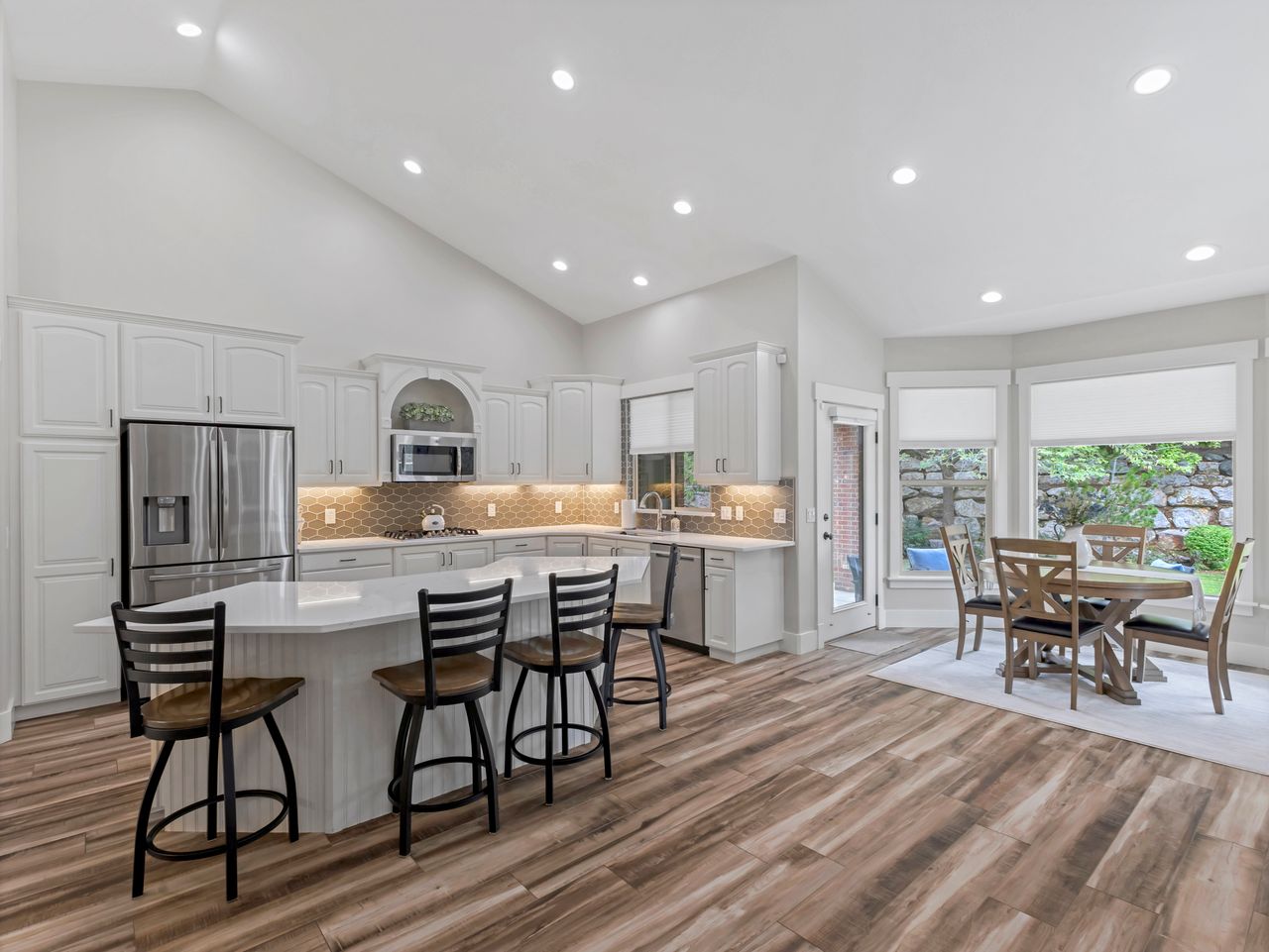 Vaulted ceiling kitchen featuring white cabinetry, stainless steel appliances, a large island with seating, wood-finish flooring, and a dining area with multiple windows