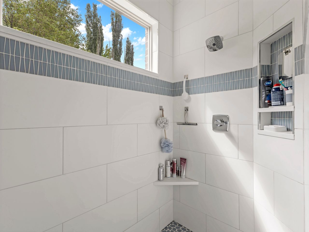 Tiled shower enclosure featuring white subway tile, a blue decorative accent band, a built-in shower niche, a corner shelf, and a window with natural light