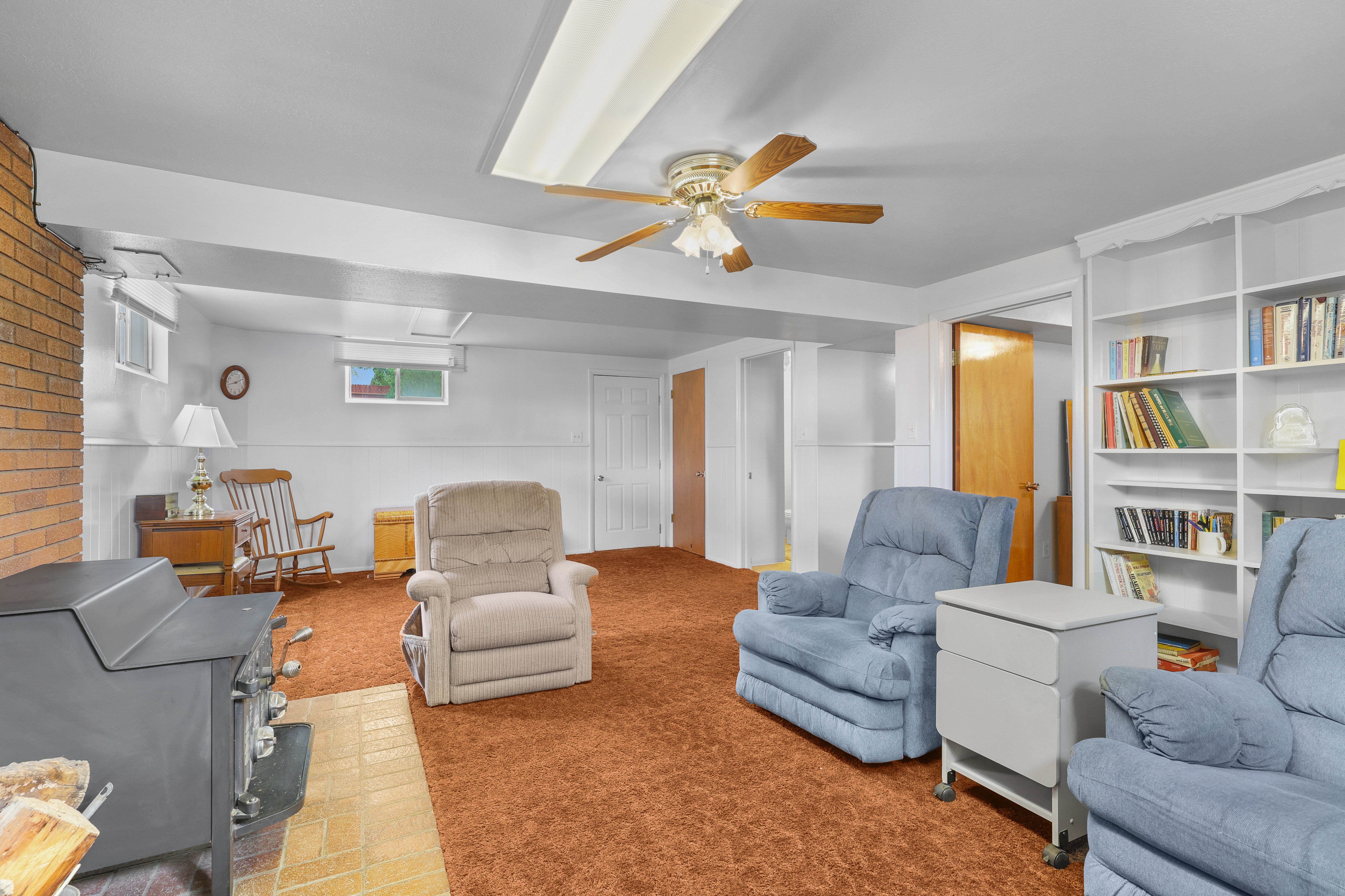 Spacious room featuring a brick-clad wood-burning stove with a tile hearth, built-in shelving, carpeted flooring, and a ceiling fan