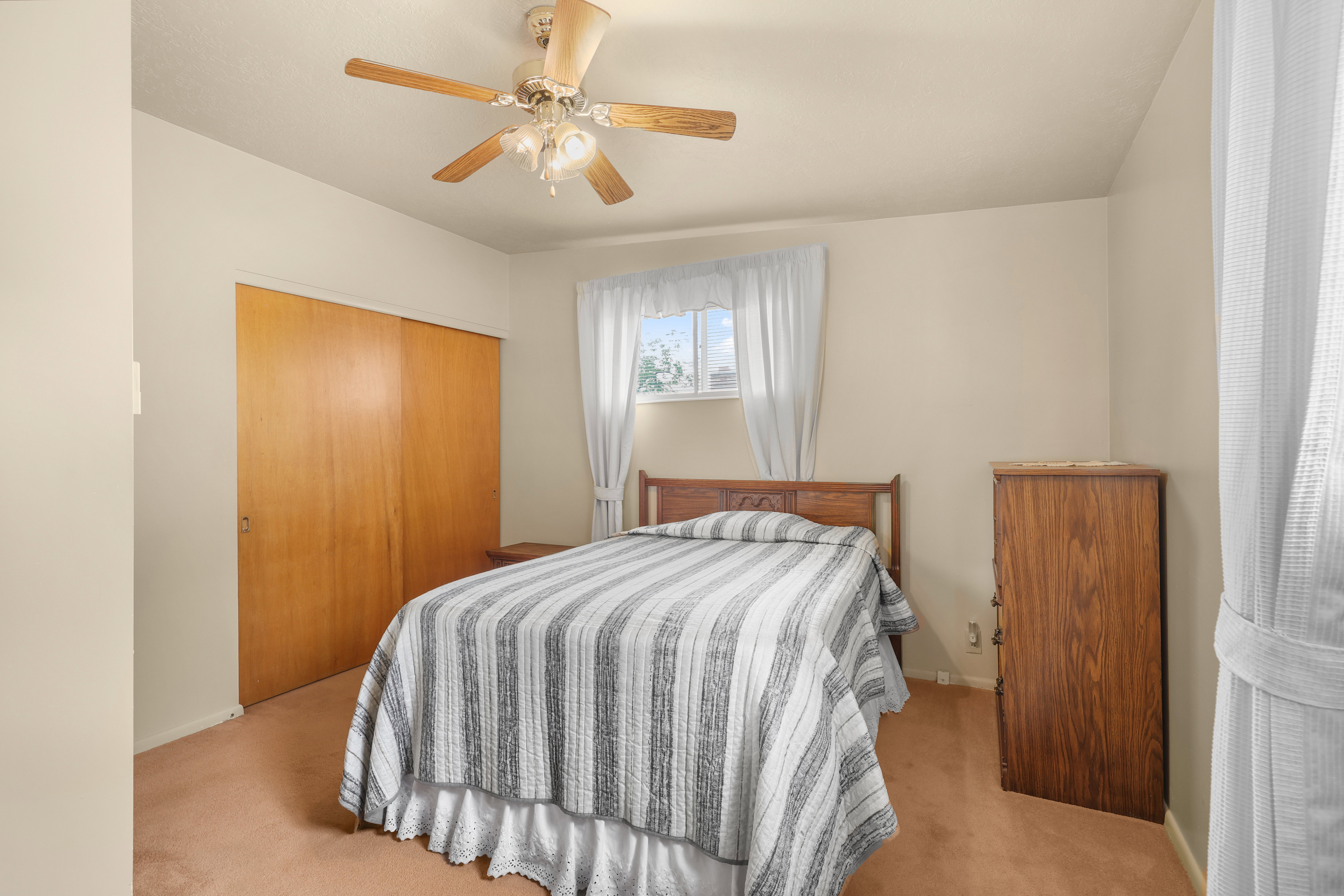 Bedroom featuring wood-finish closet doors, carpet flooring, a window with sheer curtains, and a ceiling fan with integrated lighting