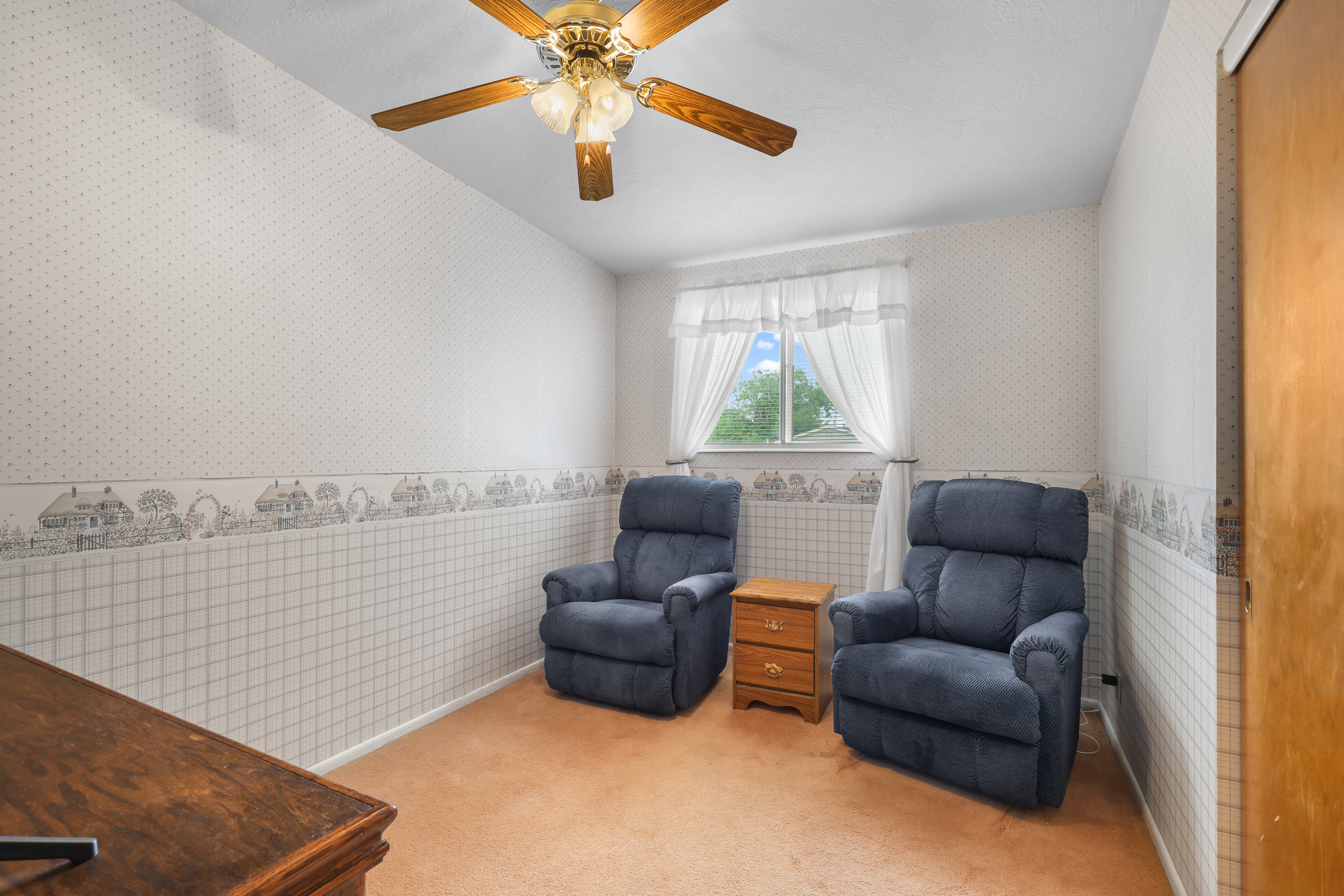 Carpeted room featuring a ceiling fan with integrated lighting, a window with sheer curtains, and light-toned wallpaper