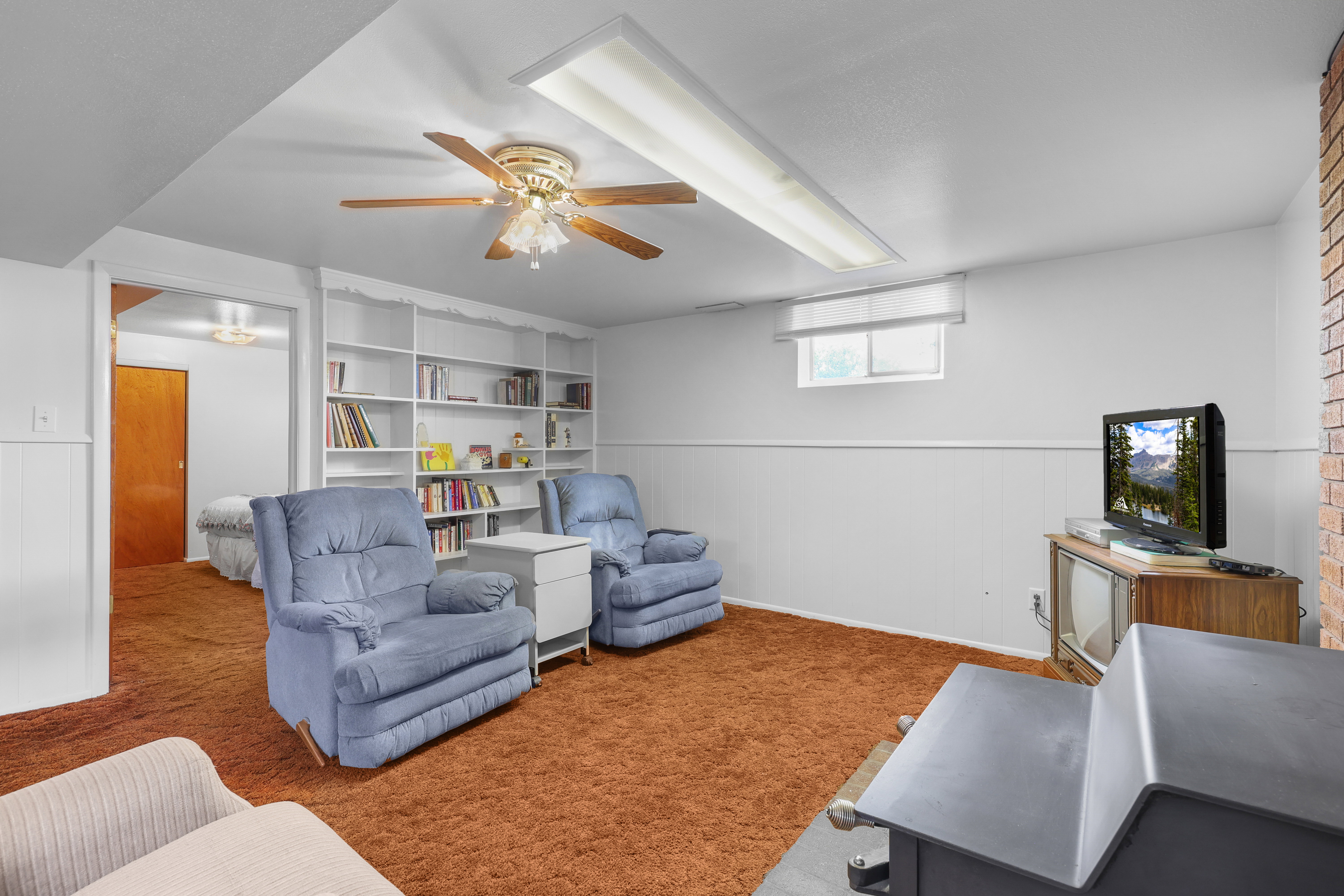 Lower level recreation area featuring wall-to-wall carpeting, built-in shelving, a ceiling fan with light fixture, a window with window treatment, and brick detailing