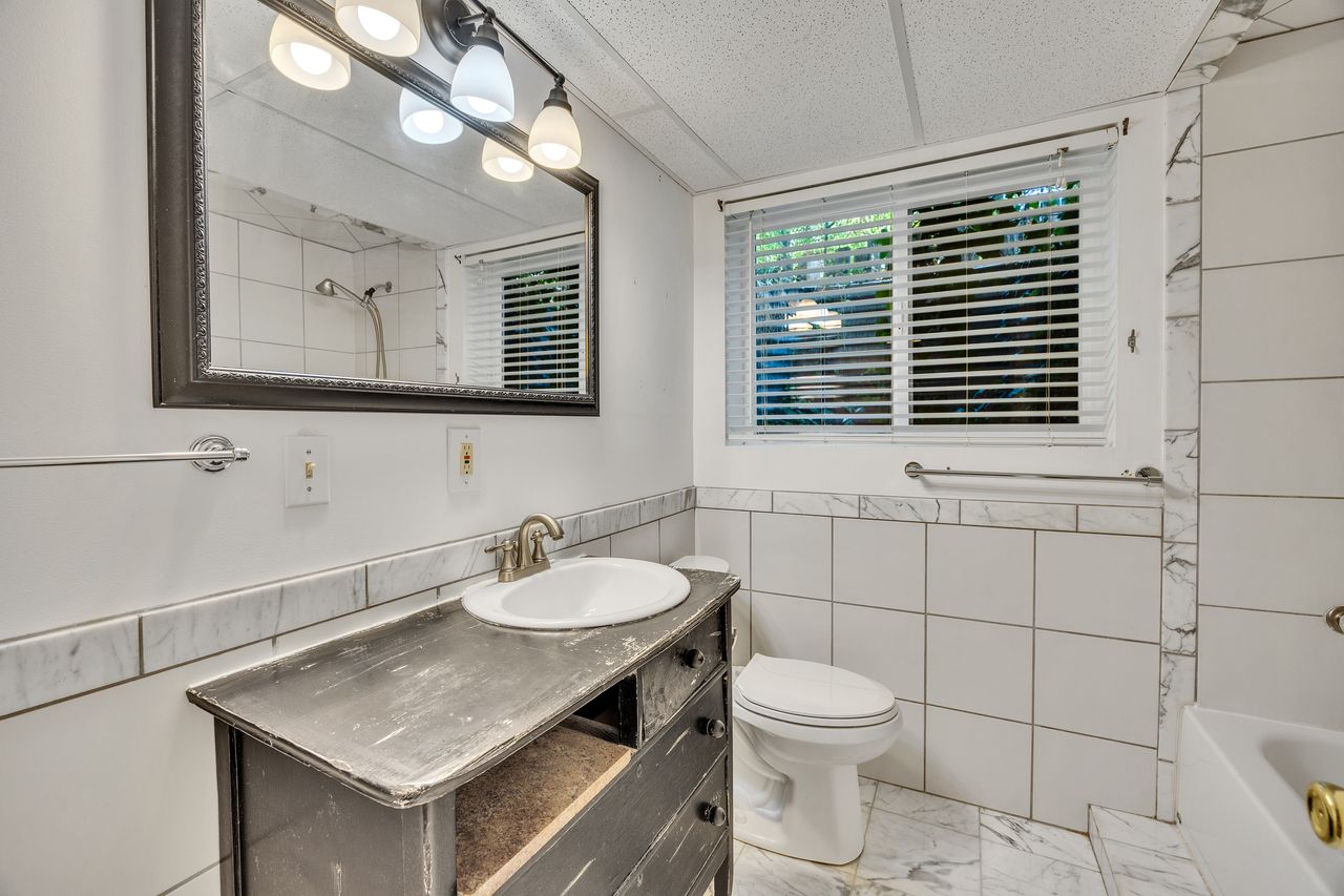Bathroom featuring a dark wood-finish vanity with a drop-in sink, white tiled walls with decorative marble-look accents, a white toilet, and a window with horizontal blinds
