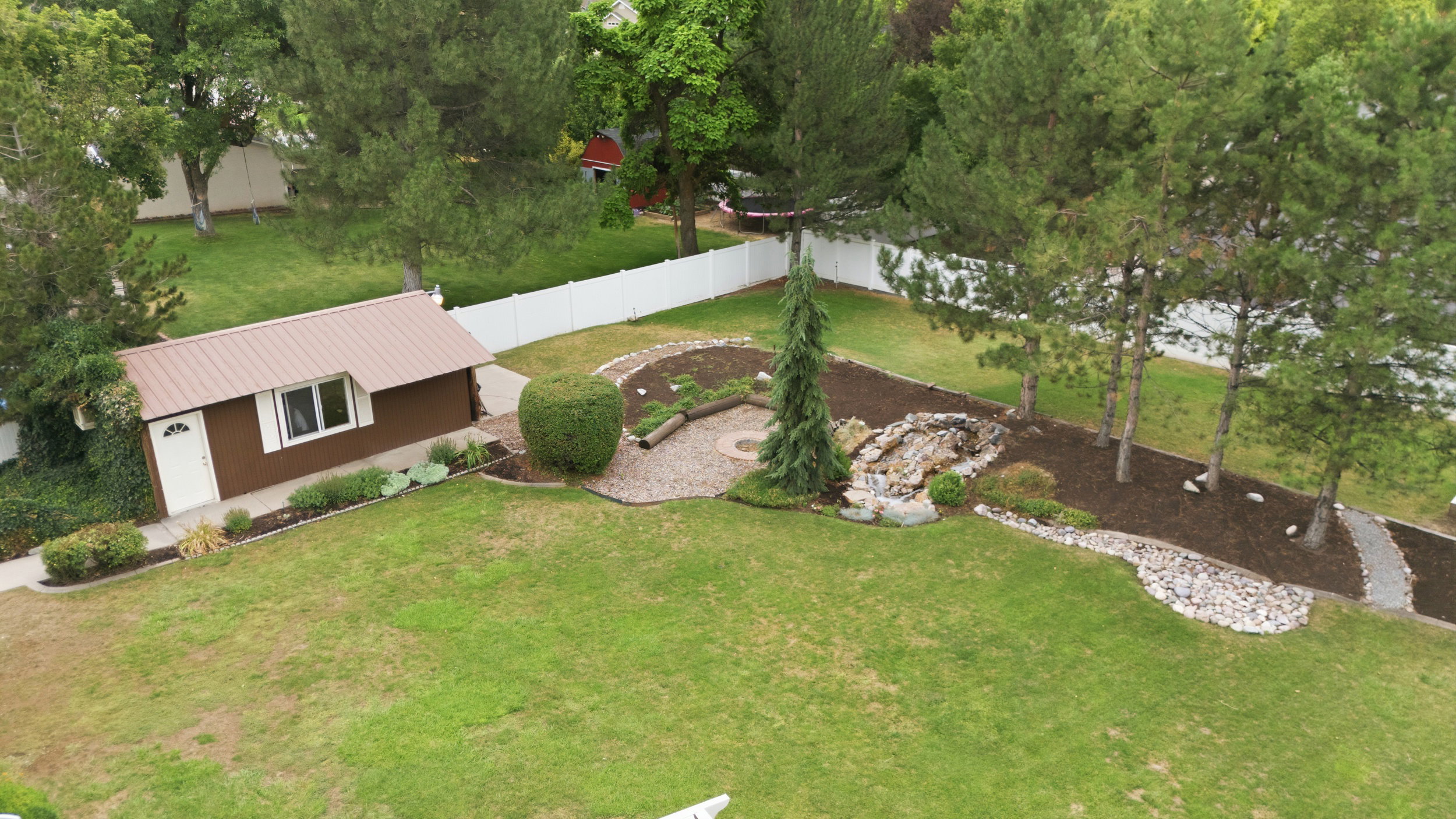 Expansive green lawn featuring a detached brown accessory building with a metal roof, a decorative rock waterfall, mature evergreen trees, and a white picket fence