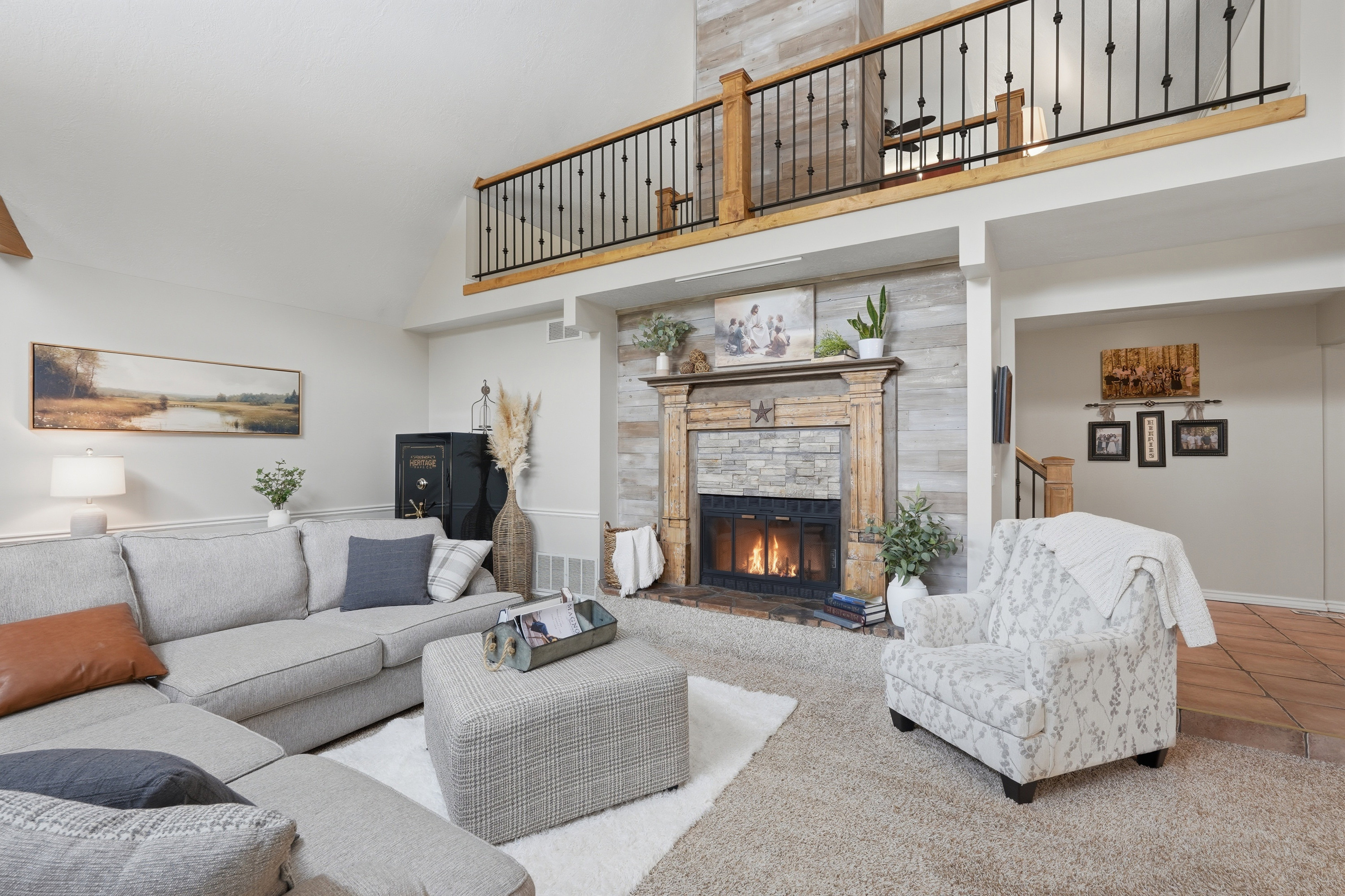 Vaulted ceiling living room featuring a stone-faced fireplace with a wood mantel, a second-story interior balcony with wood and iron railings, and light-toned carpeting