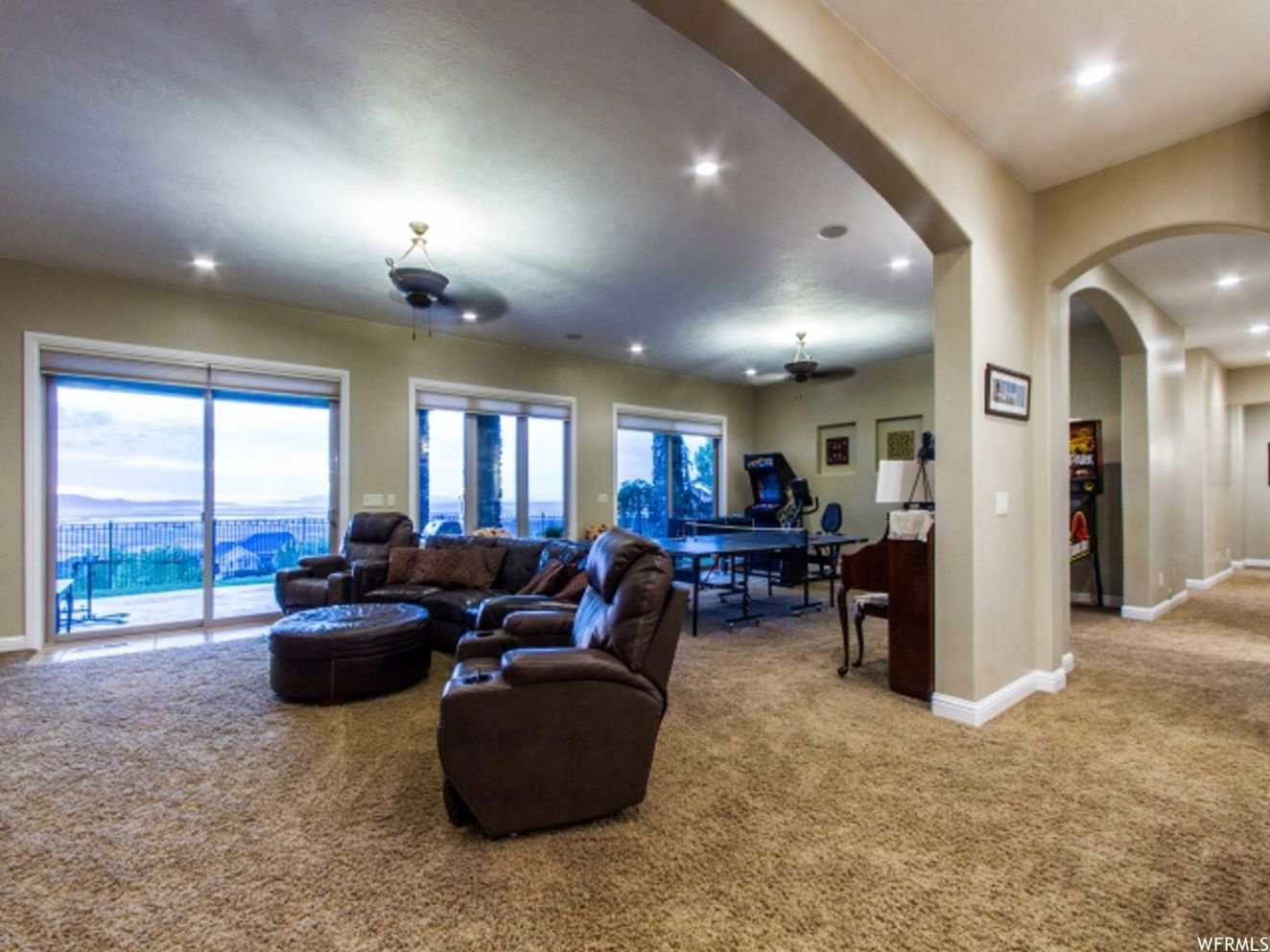 Spacious entertainment room featuring recessed lighting, plush carpet flooring, multiple sliding glass doors, ceiling fans, and a wide arched entryway