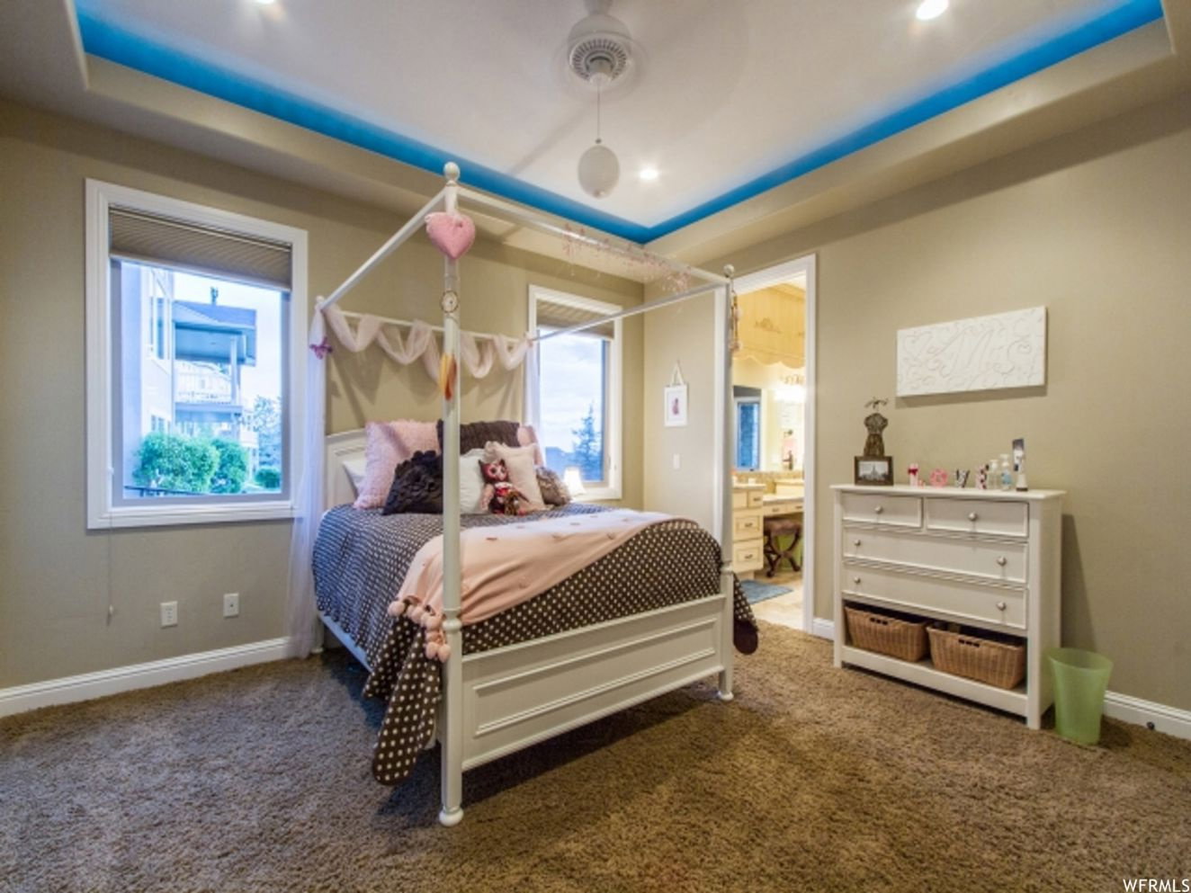 Recessed lighting and a tray ceiling with accent lighting illuminate this carpeted room, featuring white baseboards, two windows with blinds, and a doorway opening to a bathroom with a vanity