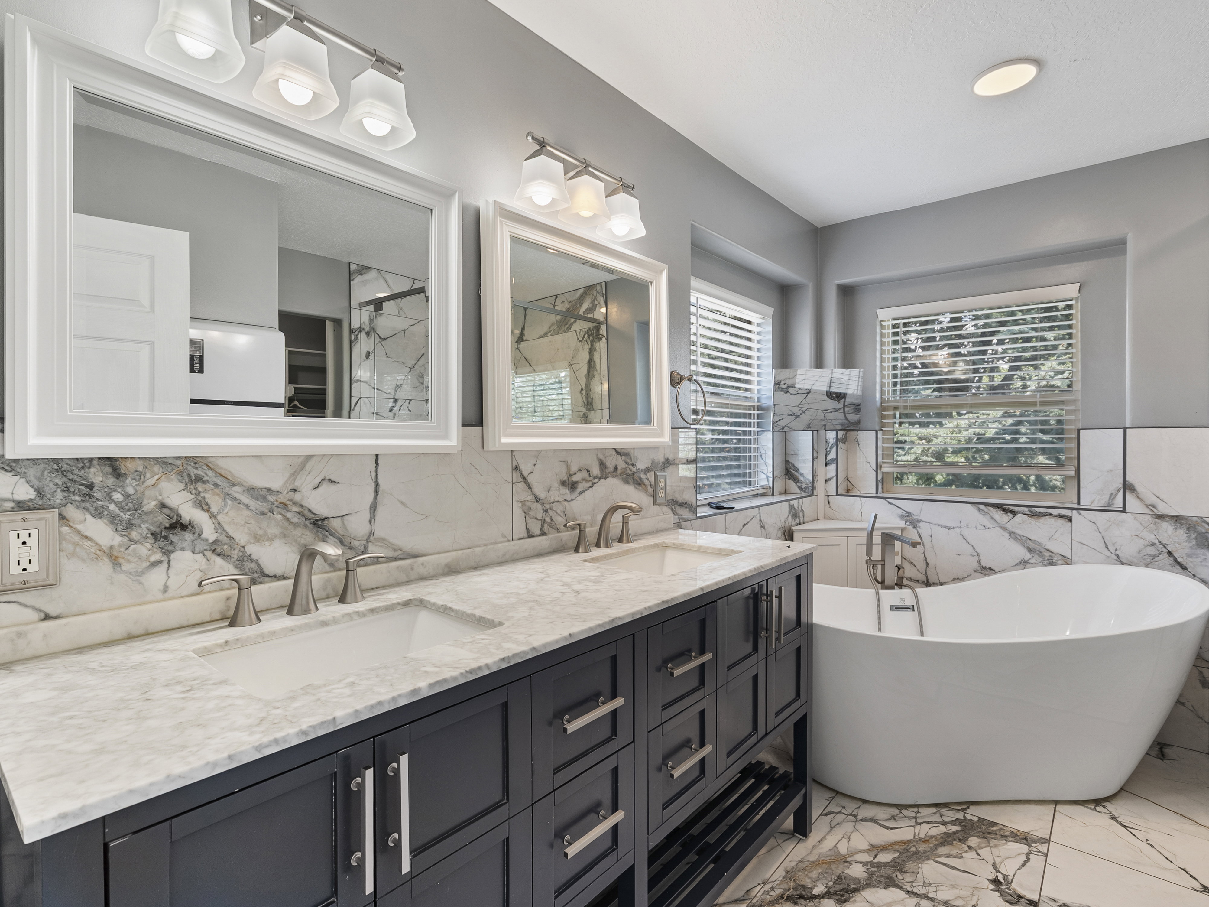 Expansive bathroom featuring a double vanity with stone countertops and undermount sinks, a freestanding soaking tub, and floor-to-ceiling marble-look tile