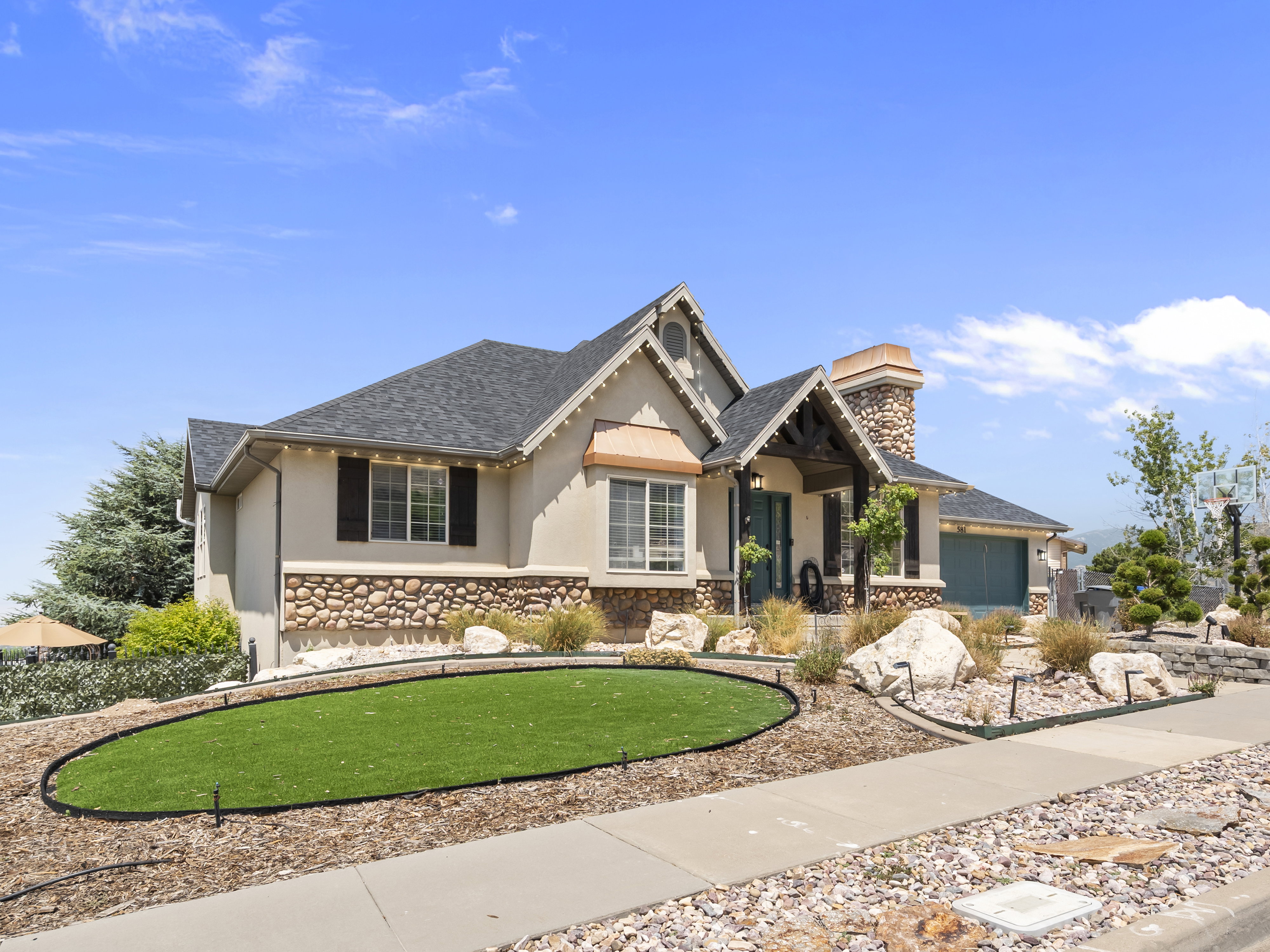 Multi-level stucco exterior with stone accents, featuring a prominent stone chimney, dark shake-style roof, and decorative window shutters