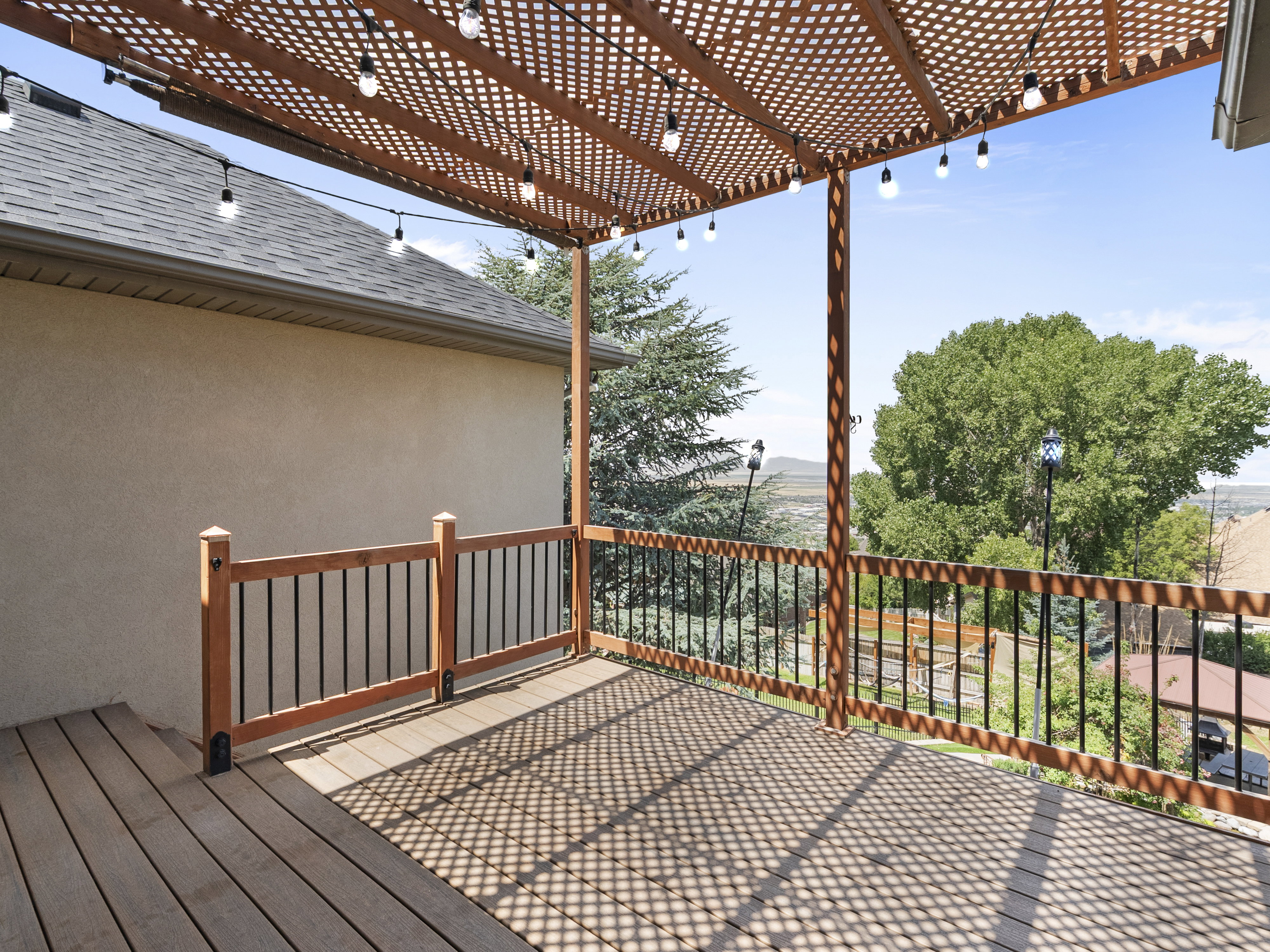 Expansive outdoor deck featuring wood-finish flooring, a lattice-style pergola with string lighting, and wood-finish railings with black balusters