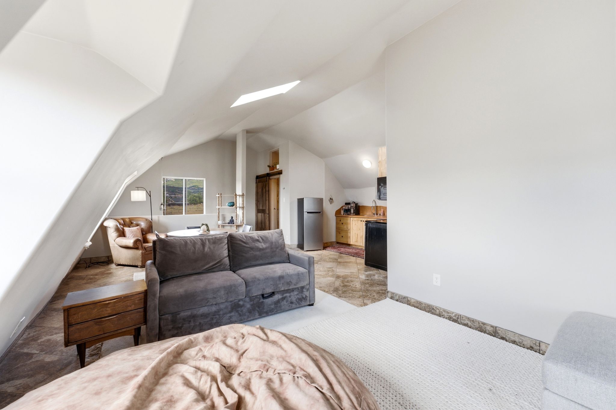 Open-concept living area featuring a vaulted ceiling with skylight, a window, wood-finish flooring, and a compact kitchen area with wooden cabinetry and a built-in refrigerator