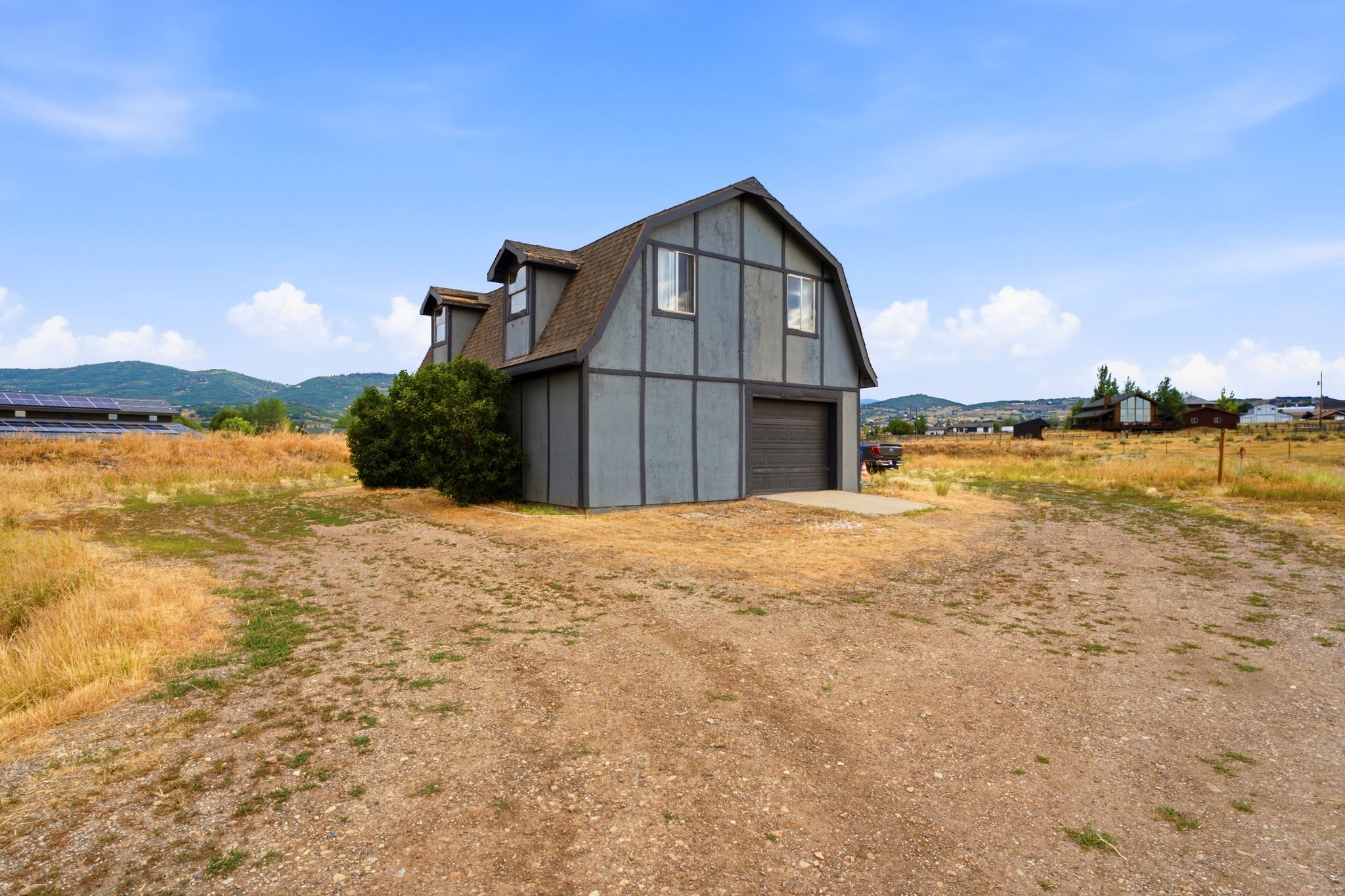 Two-story structure with Gambrel roofline, featuring dormer windows, a two-car garage, and a gravel driveway