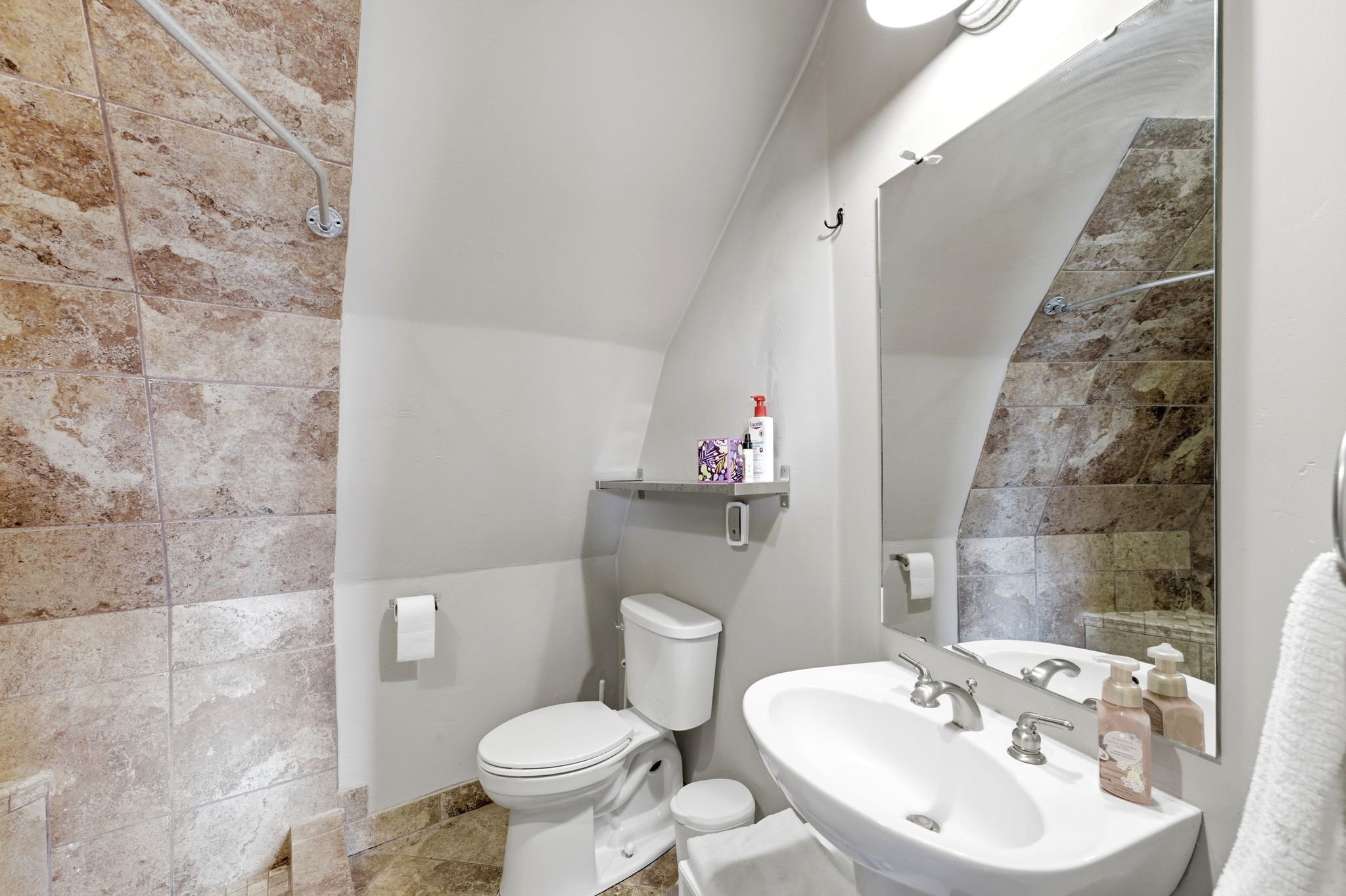 Bathroom featuring a wall-mounted sink, a toilet, a decorative wall mirror, and extensive natural stone-look tilework on the shower surround and flooring