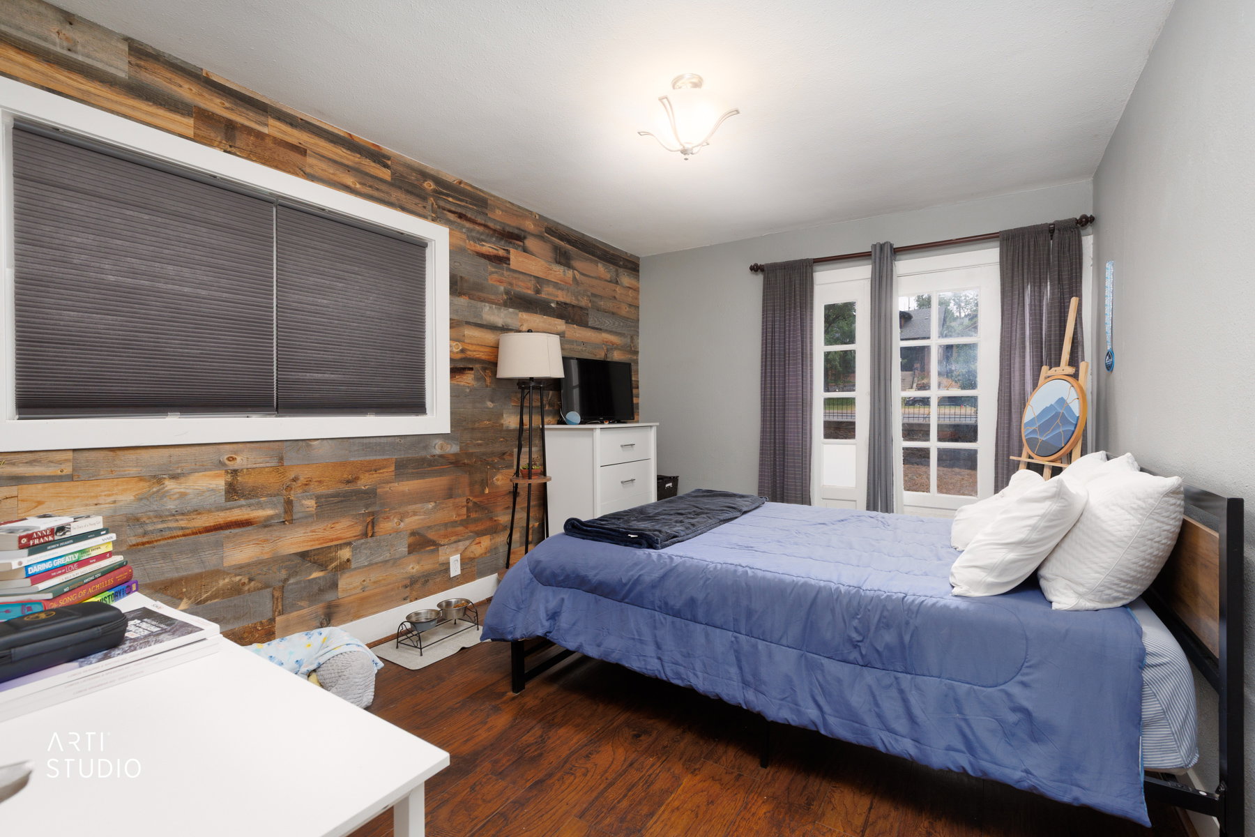 Bedroom featuring a prominent wood-panel accent wall, wood-finish flooring, a white window frame with blinds, a white ceiling, and a double glass-panel door with privacy curtains