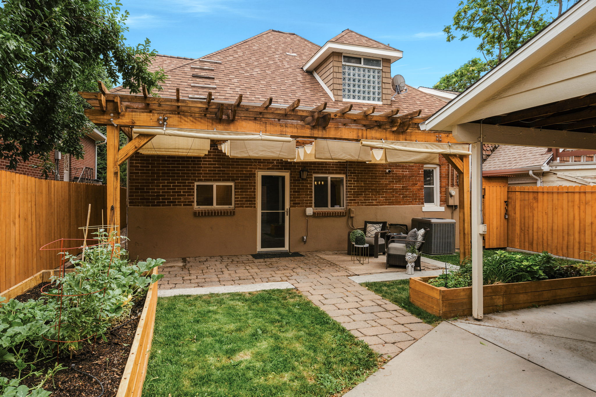Rear exterior featuring a brick facade with a built-in pergola, paver patio, established lawn, raised garden beds, and wood fencing