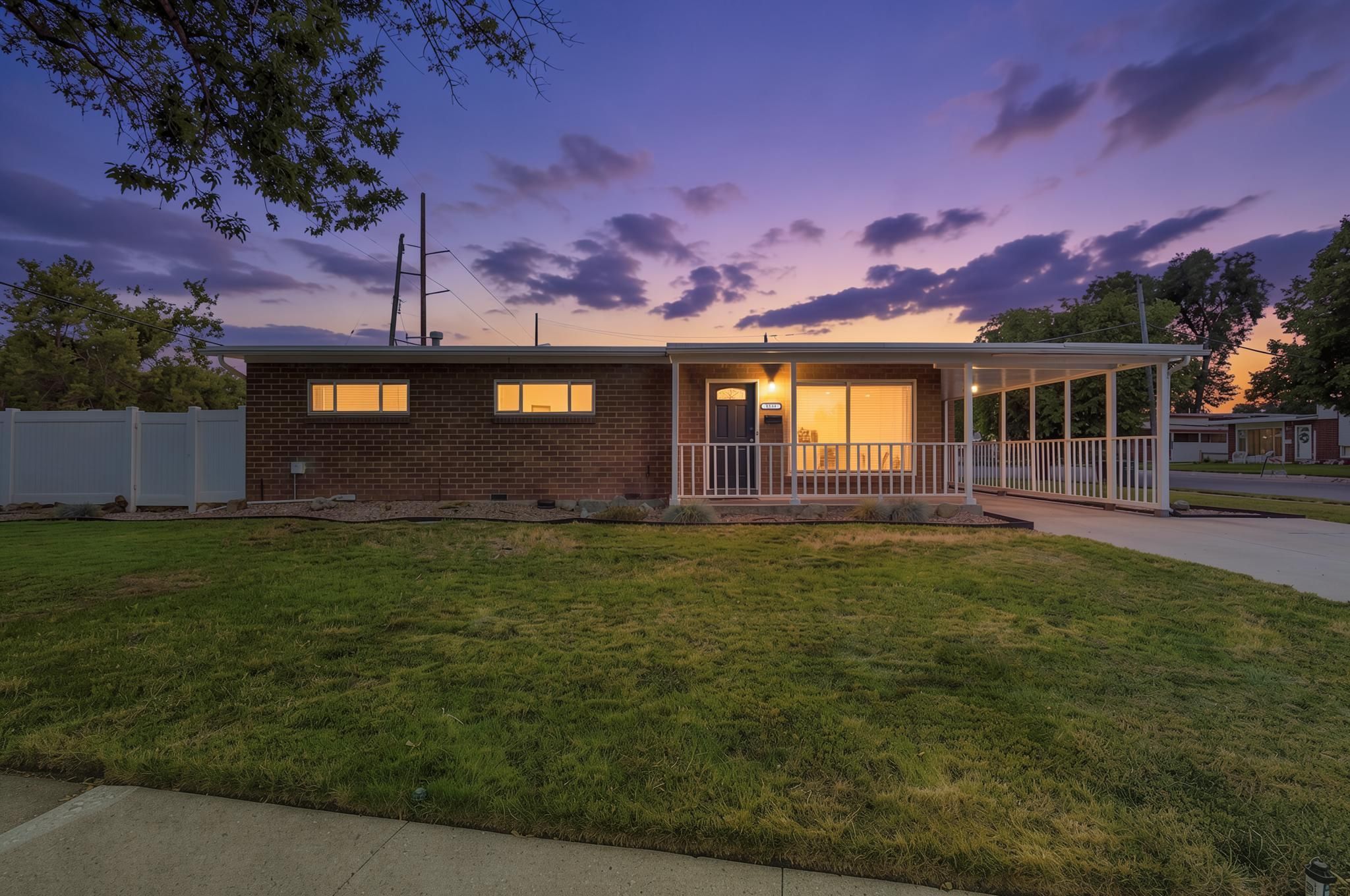 Brick exterior with a covered front porch featuring white railings, a covered carport, and a front lawn with mature trees and a white privacy fence