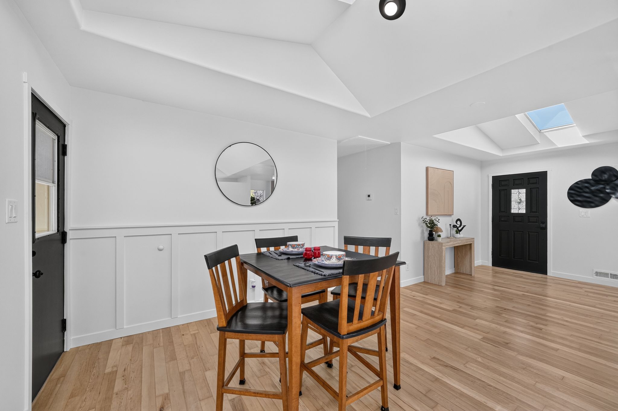 Open-concept dining area featuring light wood-finish flooring, white paneled wainscoting, and a vaulted ceiling with recessed lighting