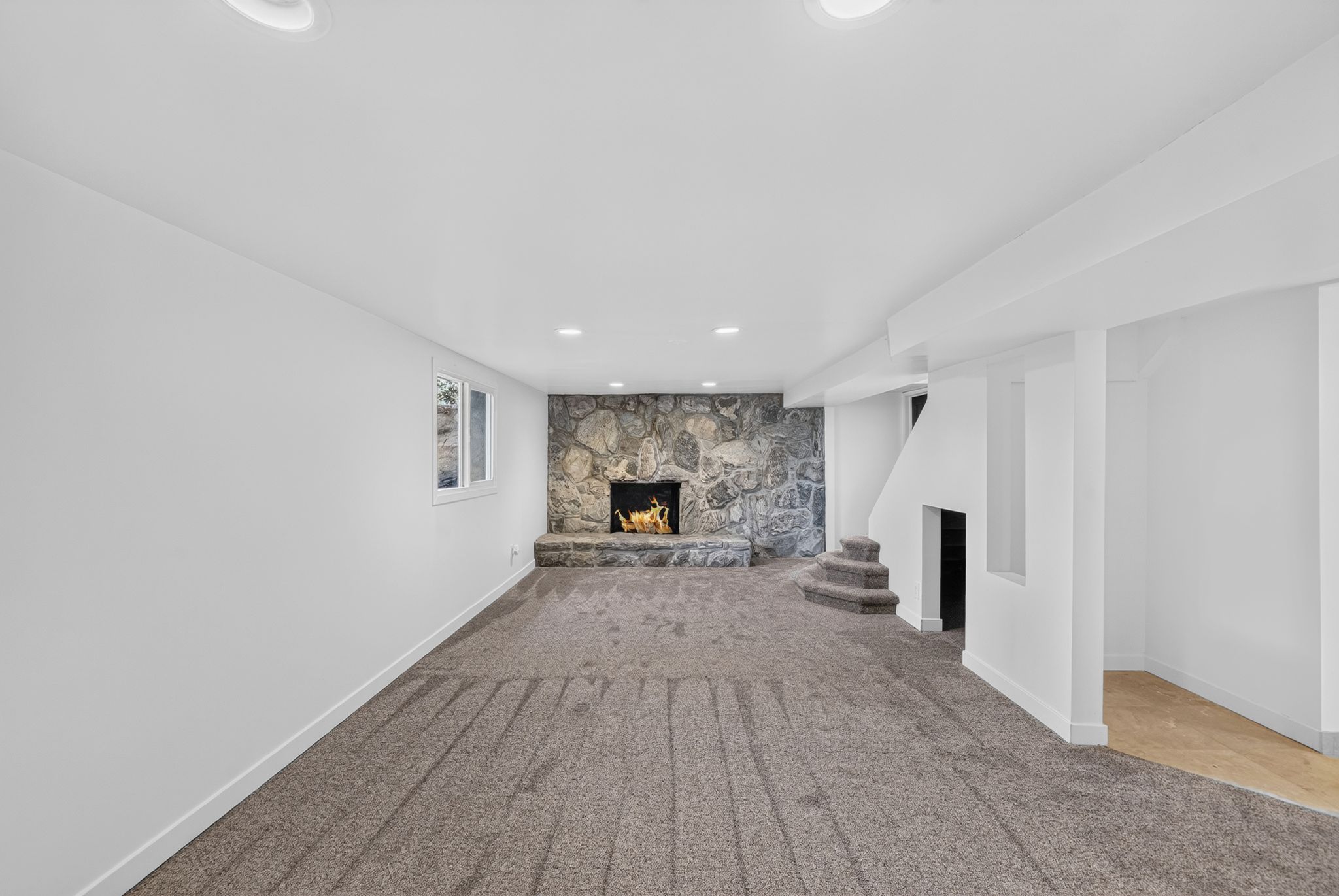 Spacious entertainment area featuring a stone-clad fireplace with hearth, recessed lighting, neutral carpeting, and a visible window